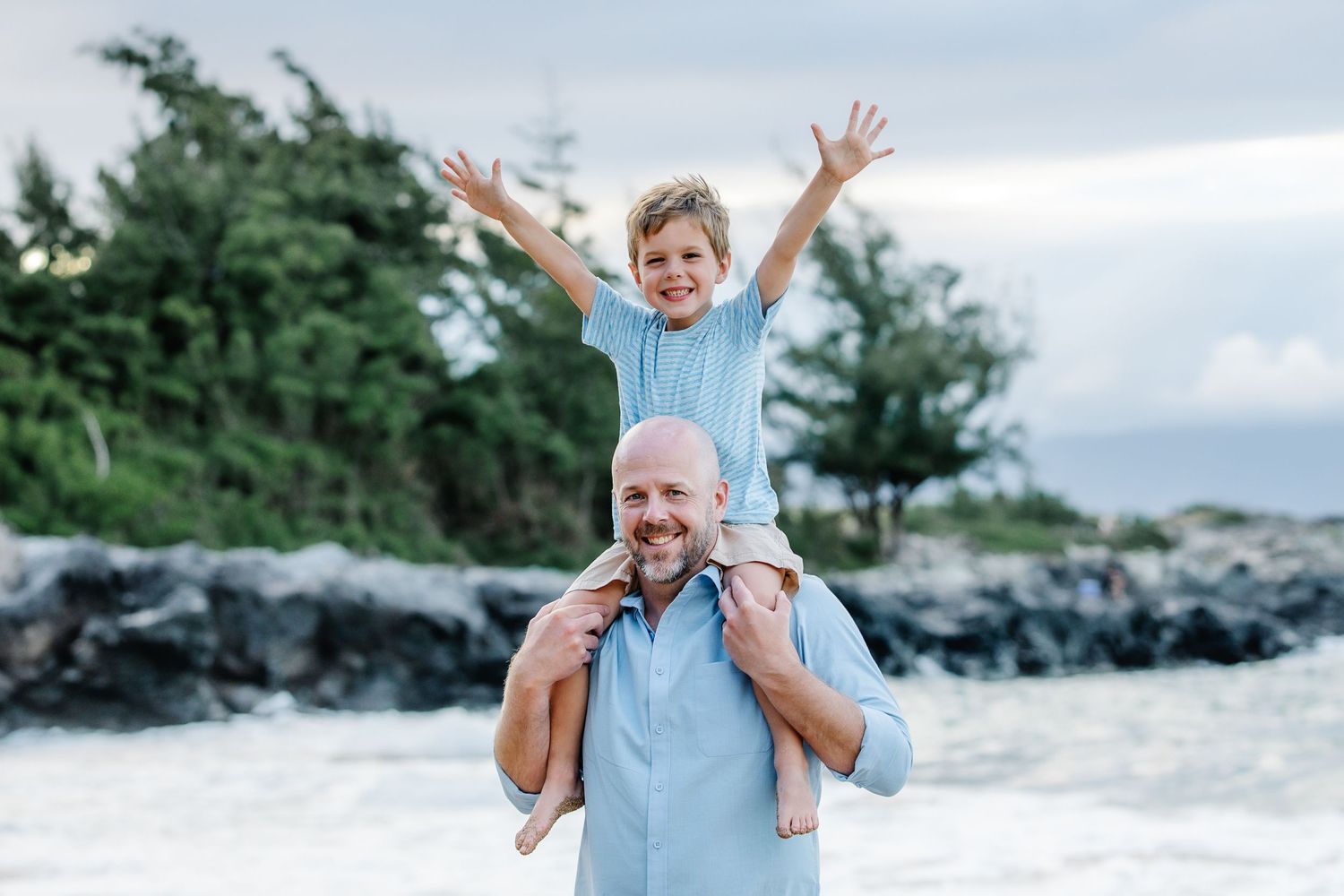 A smiling bald man carries a joyful boy on his shoulders at a rocky beach with lush green trees behind them.