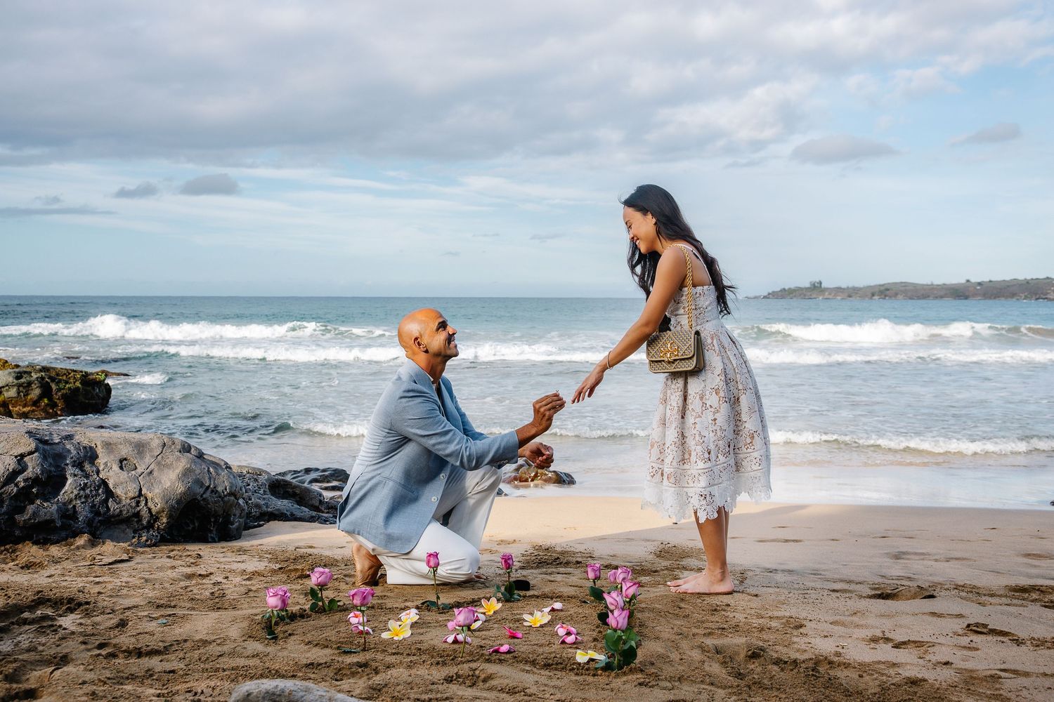 Beach proposal scene with rose petals scattered along rocky shoreline.