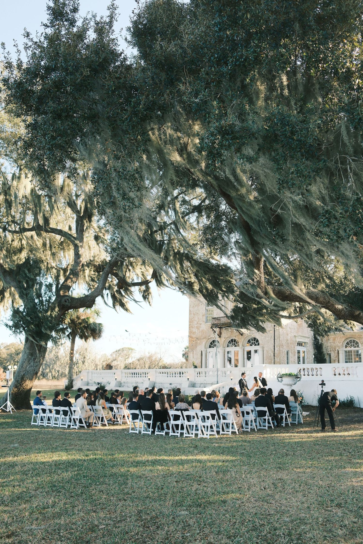 Outdoor wedding ceremony site with white chairs arranged on a lawn beneath majestic oak trees with hanging moss.