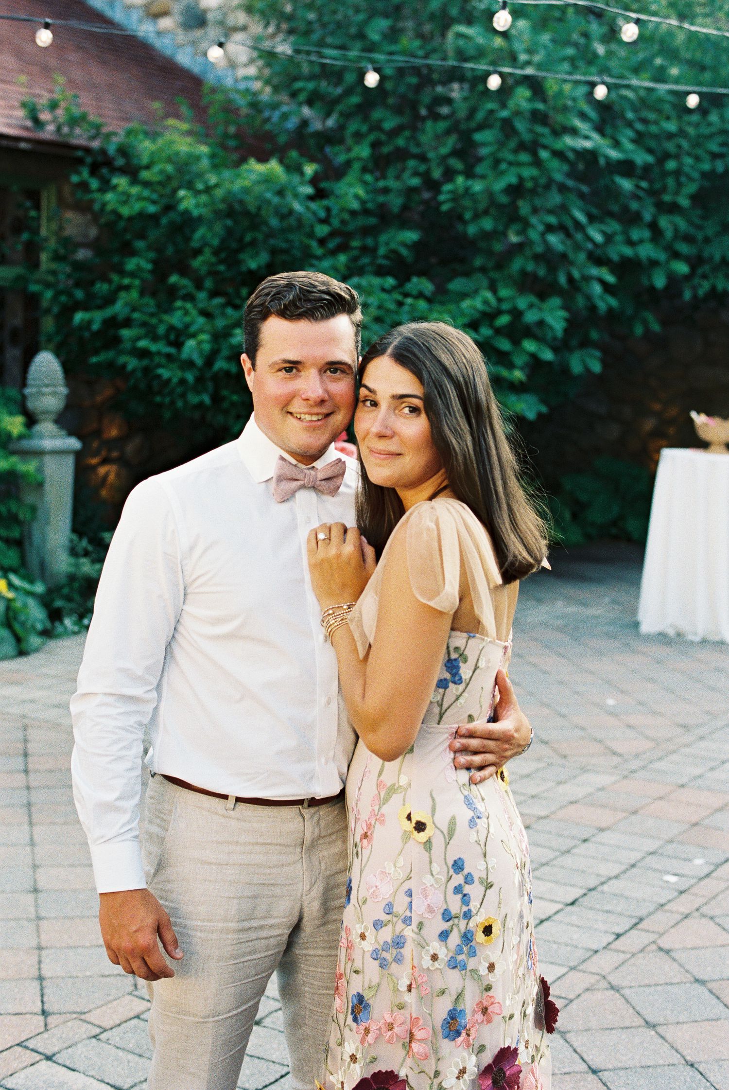 Couple posing outdoors at a garden wedding reception, man in white shirt and pink bow tie, woman in floral dress.