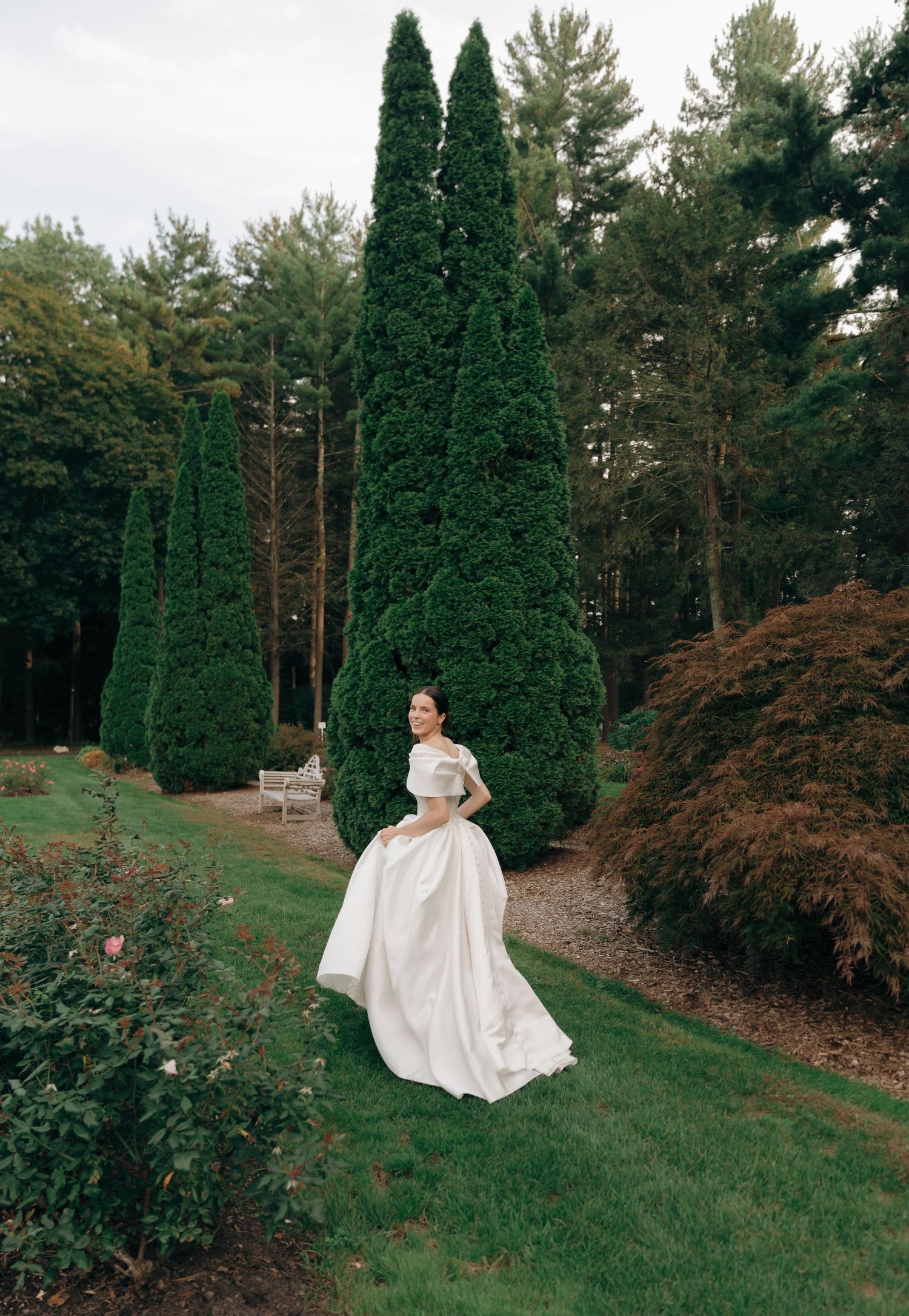A figure in a flowing white dress moves gracefully through a garden landscape with tall cypress trees and autumn foliage.