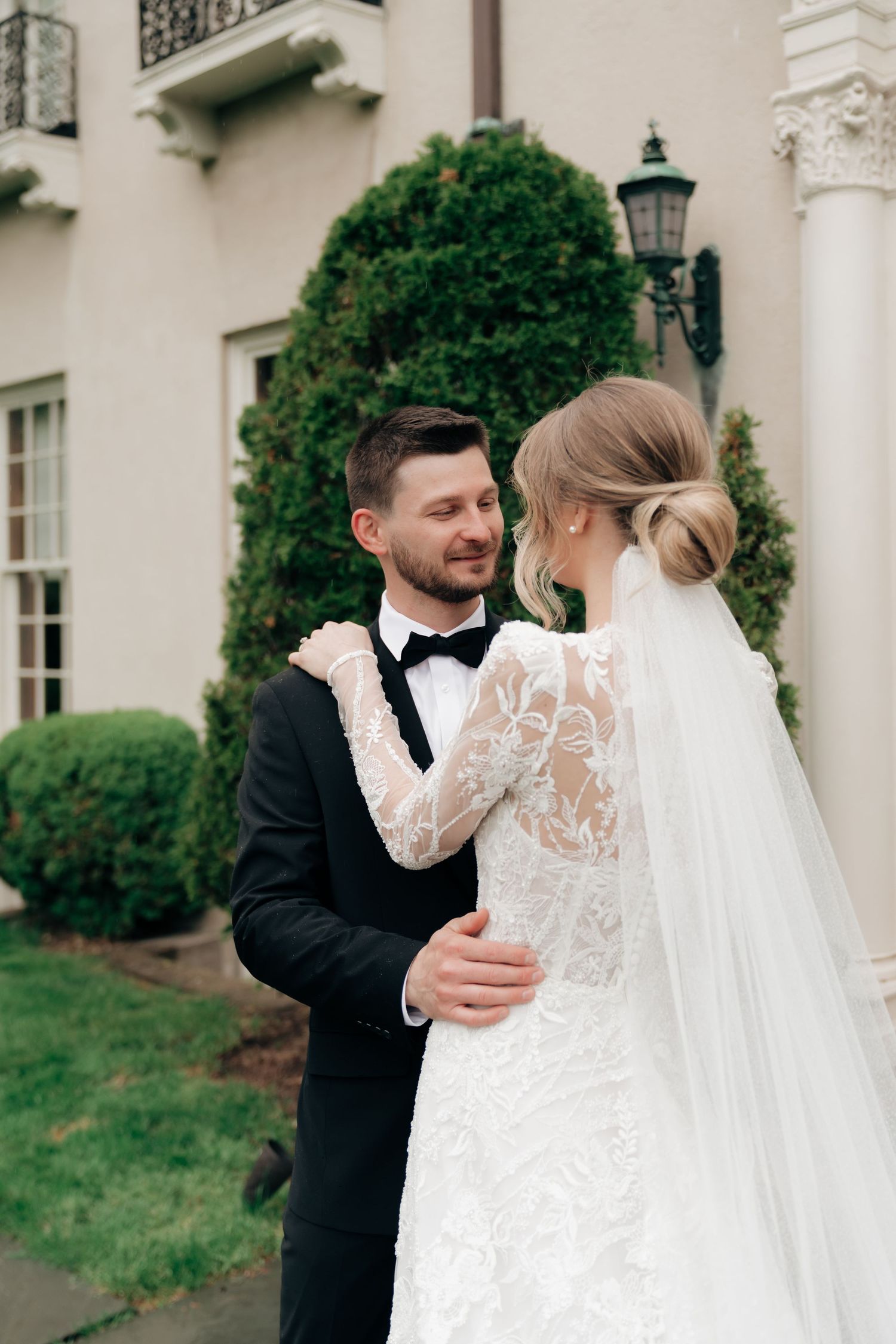 Bride in lace gown and veil embraces smiling groom in black tuxedo outside elegant white mansion.