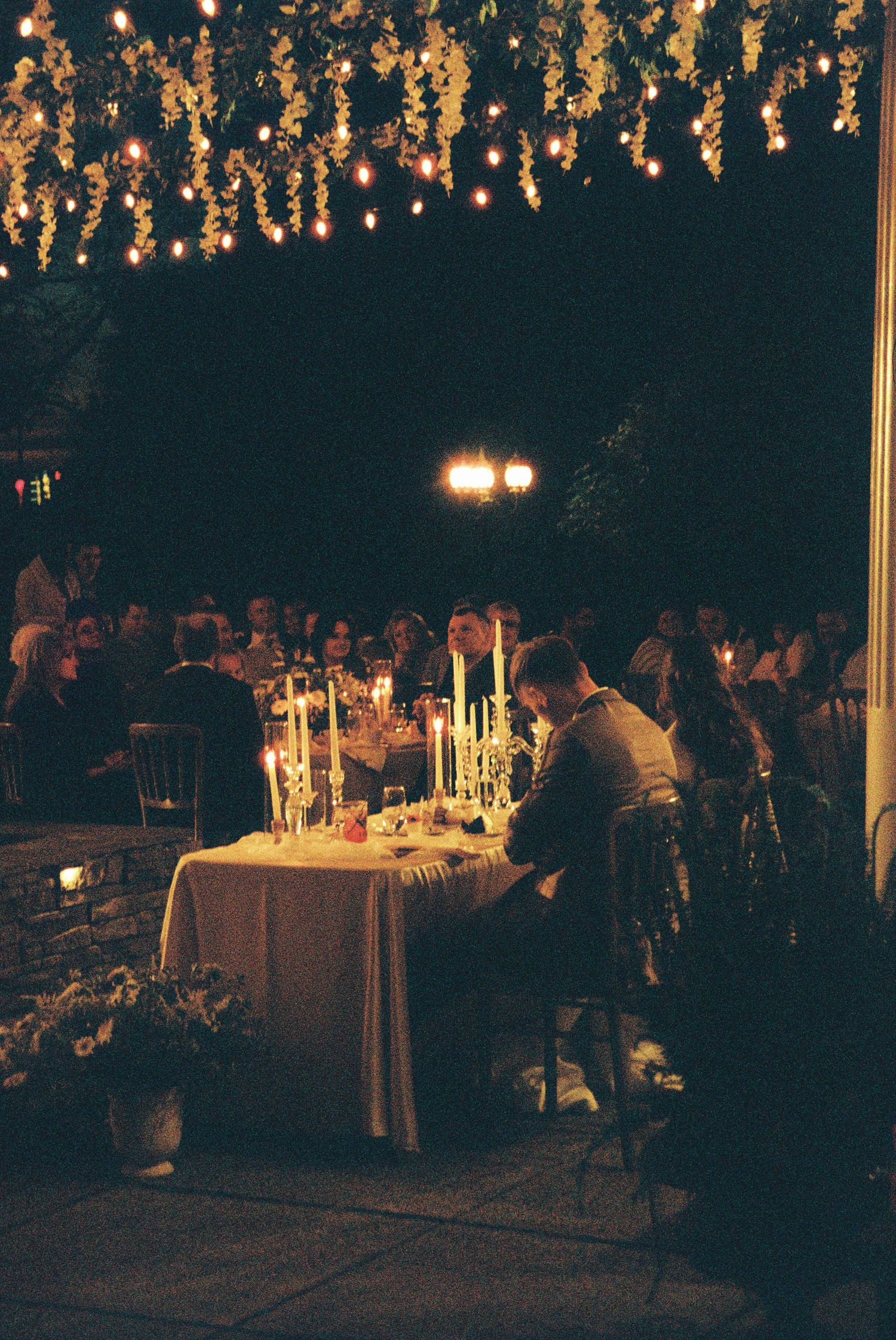 Romantic outdoor wedding reception at night with string lights, floral canopy, and candlelit sweetheart table.