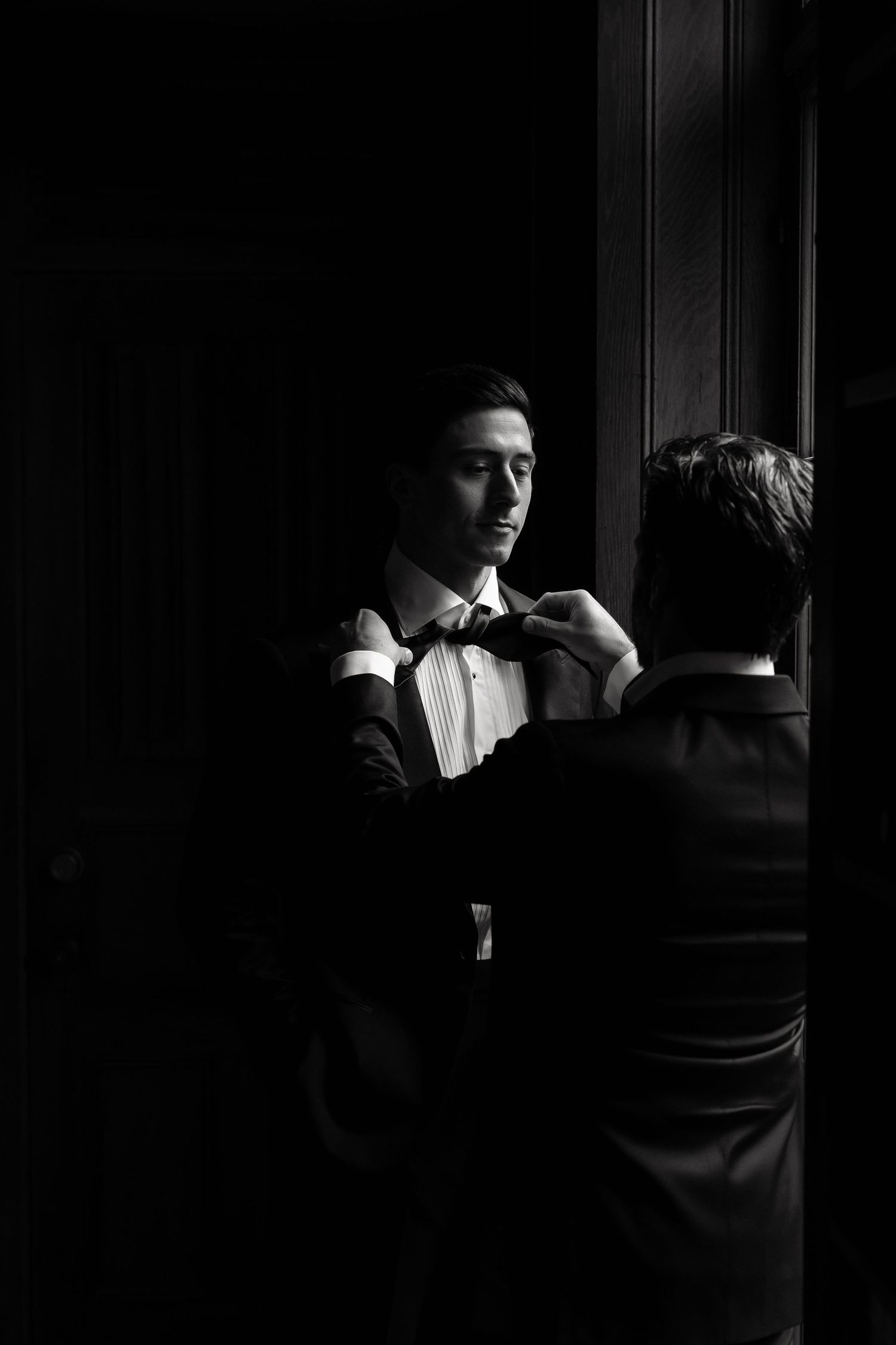 Black and white photo of groom adjusting bow tie with help from groomsman before wedding ceremony.