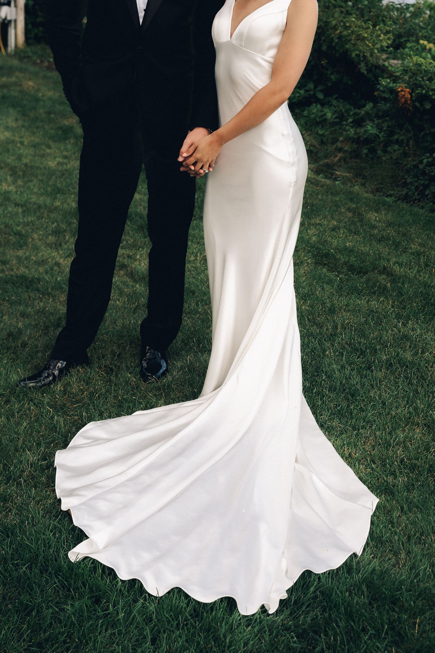 Bride in elegant white satin slip wedding gown with train holds hands with groom in black suit on green lawn.
