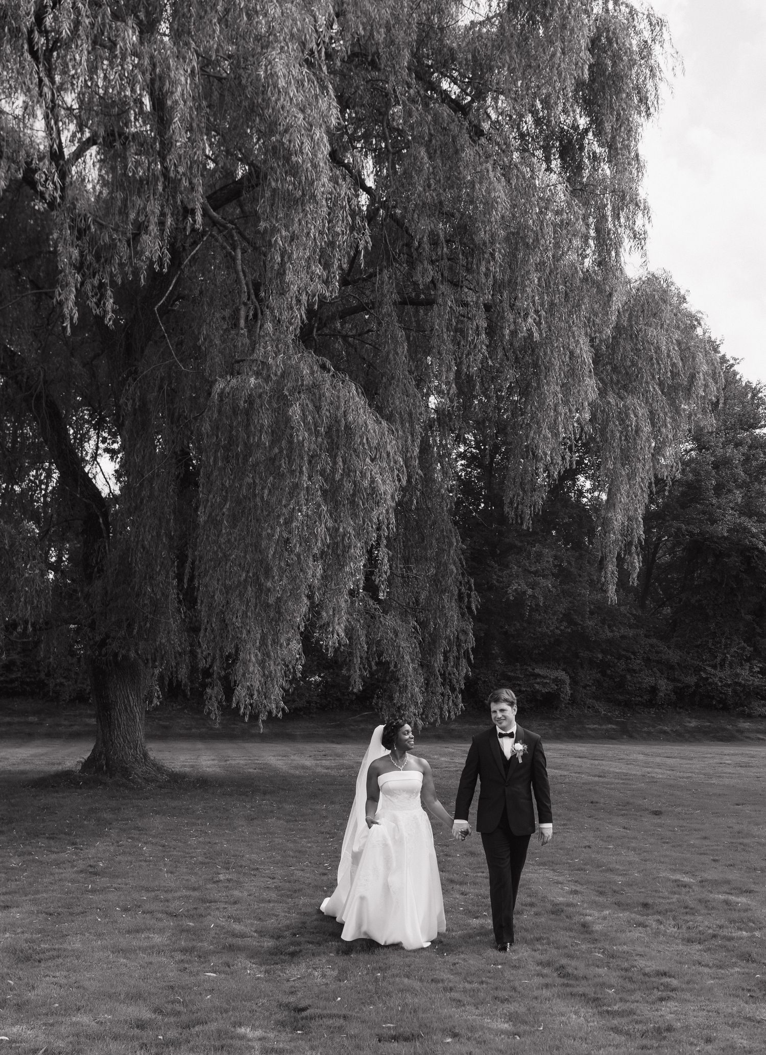 A couple in wedding attire walks together under dramatic weeping willow trees in a black and white photo.