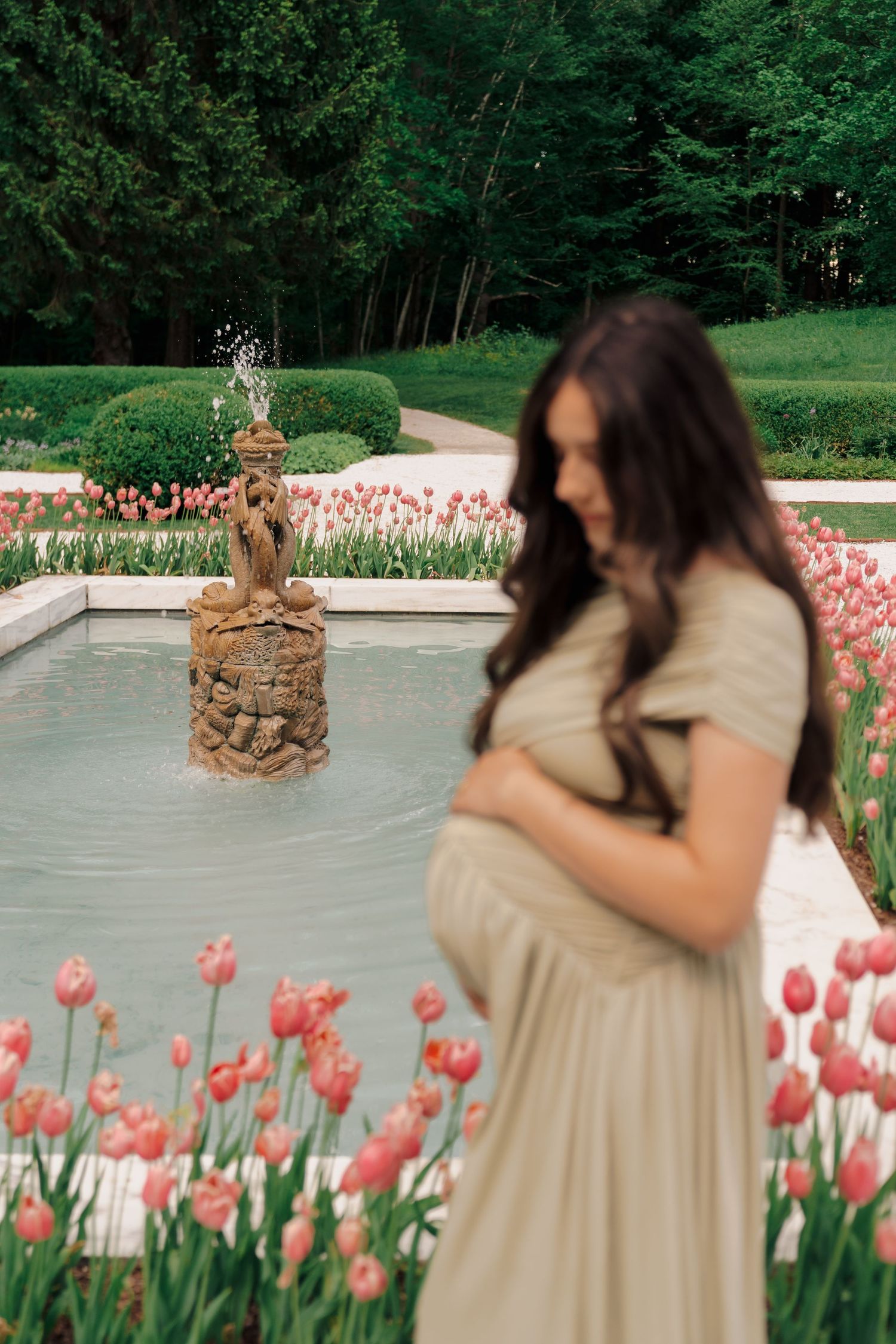 Pregnant woman in beige dress holding belly near fountain with pink tulips in lush garden park.