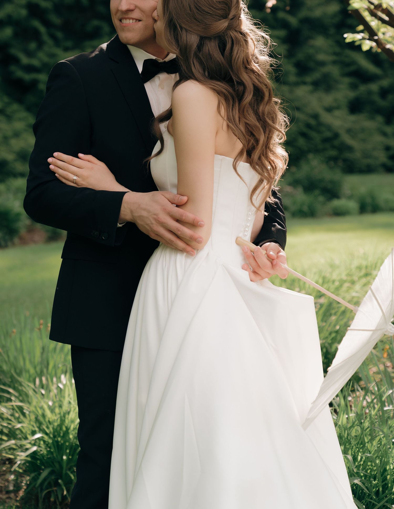 A couple in wedding attire embraces while standing in a sunlit meadow with lush green grass and trees.