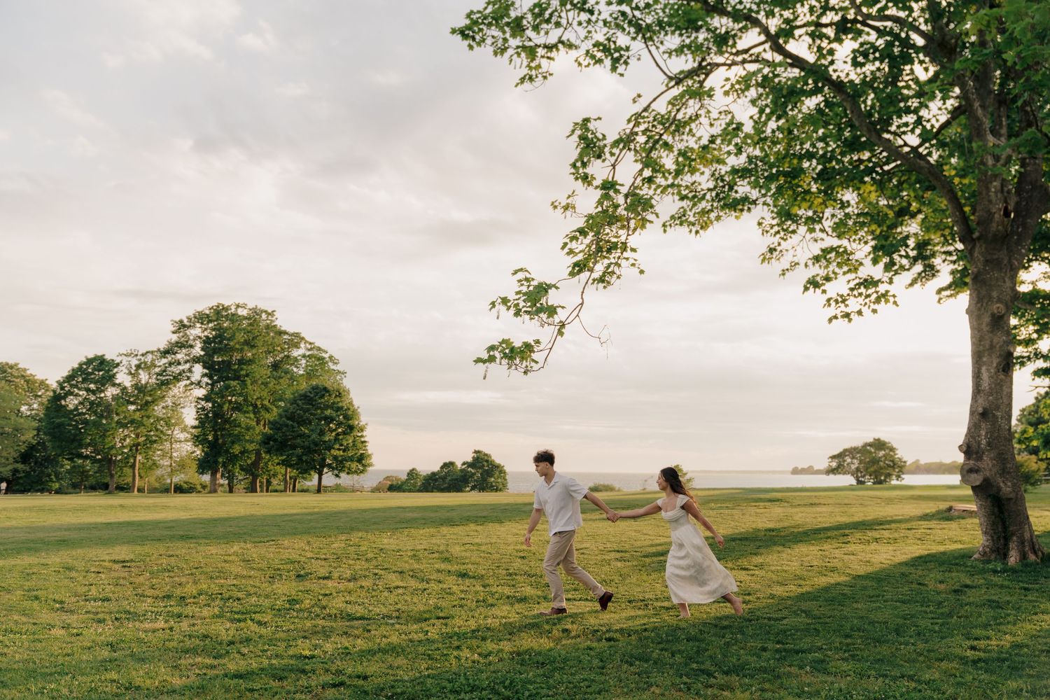 Couple holding hands running through lush green park with large trees and open sky at golden hour.