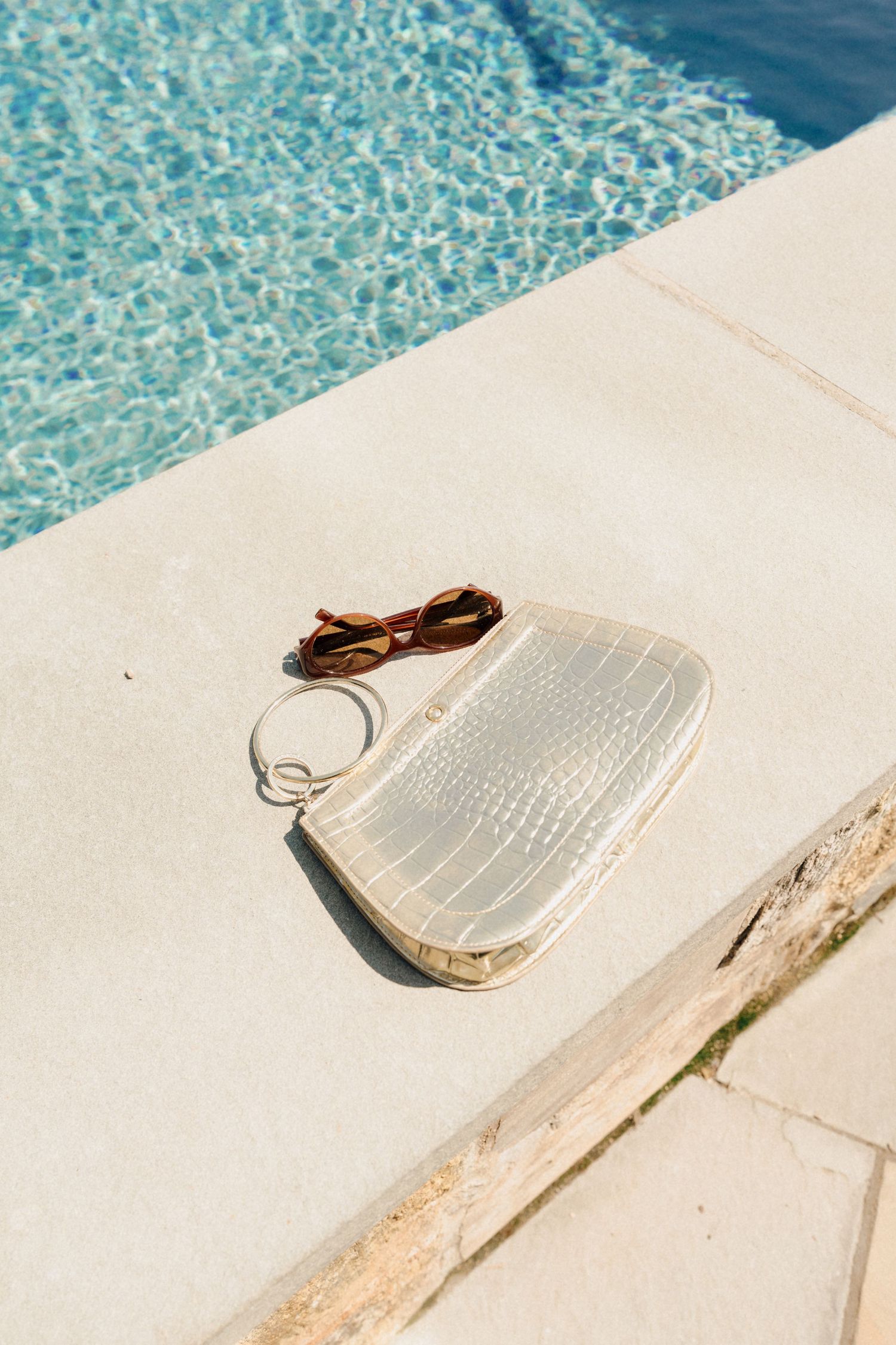 White croc-embossed clutch bag with gold ring handle and brown sunglasses on poolside edge.