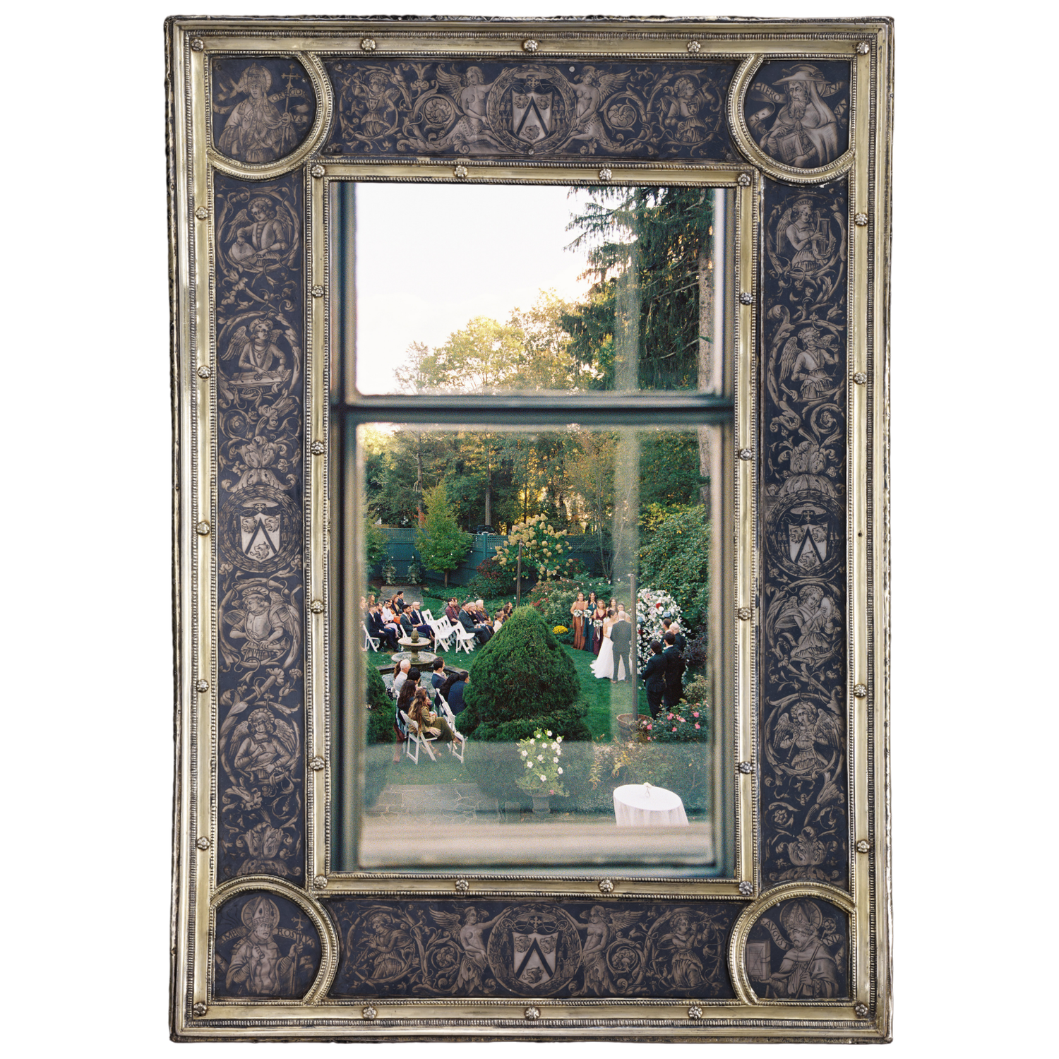 Ornate silver-framed mirror reflecting an outdoor garden wedding ceremony with guests seated among lush green trees.