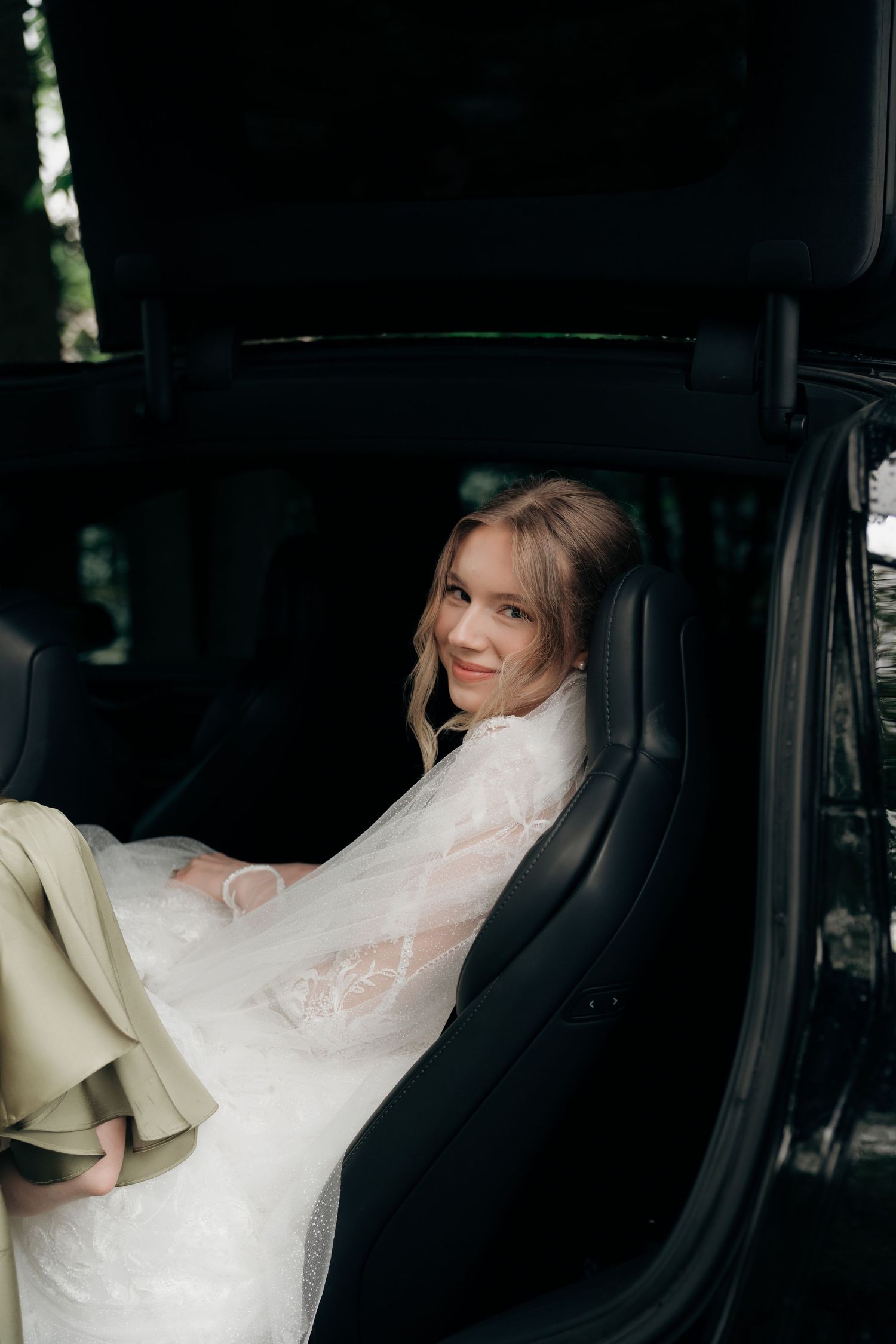 Blonde bride in lace wedding dress sitting in luxury car backseat, smiling at camera.