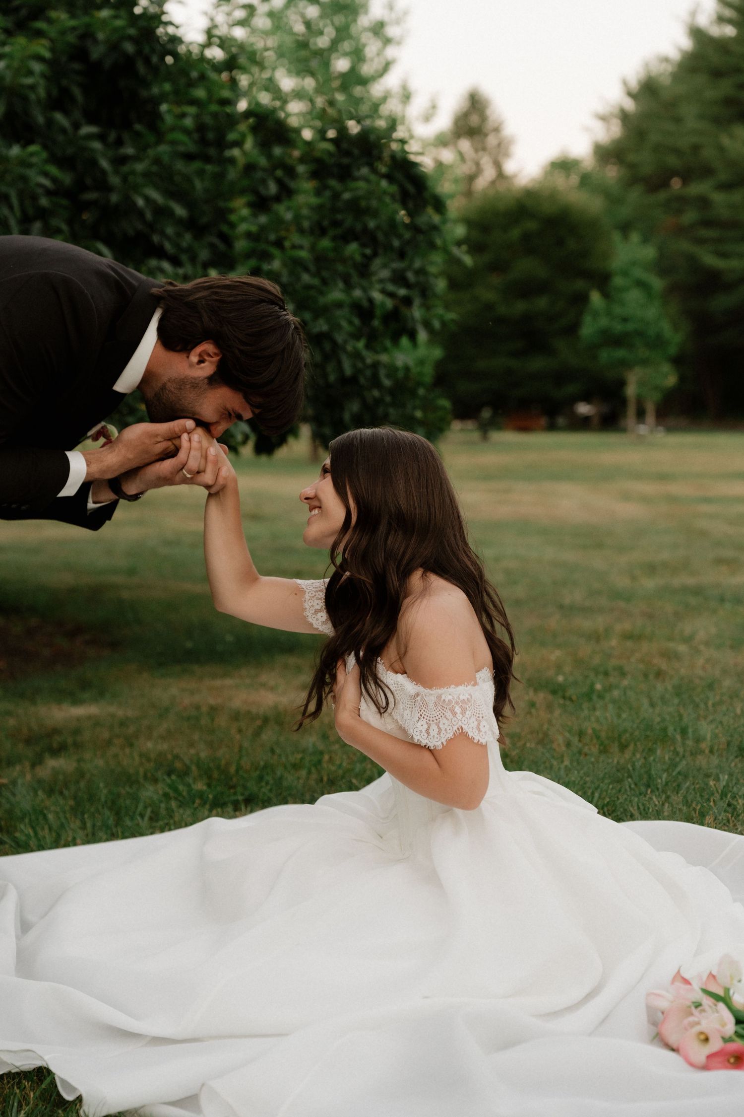 Groom kisses bride's hand as she kneels in white off-shoulder gown on green lawn during outdoor wedding.