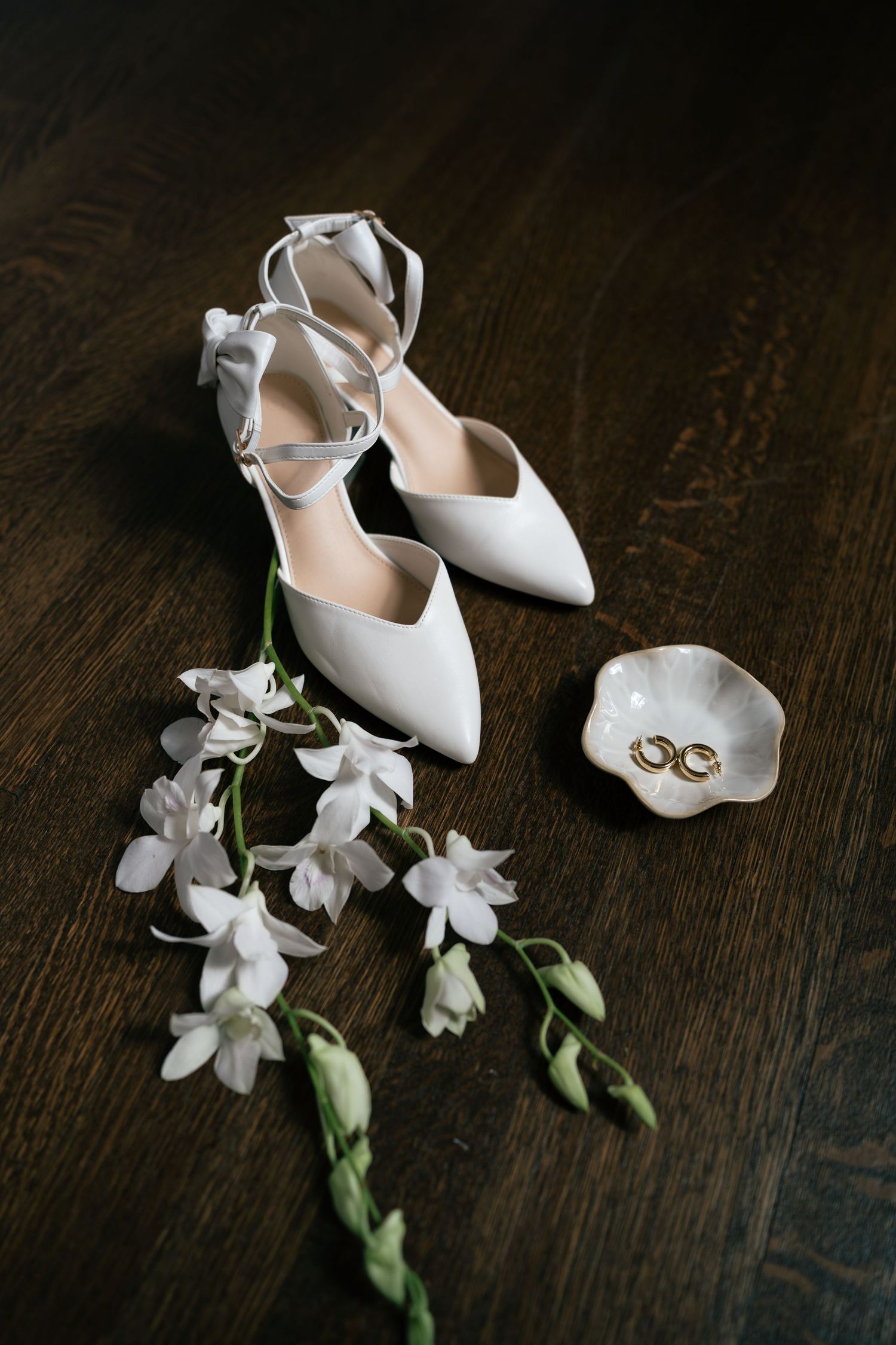 White wedding shoes and delicate white flowers arranged on a dark wooden surface.