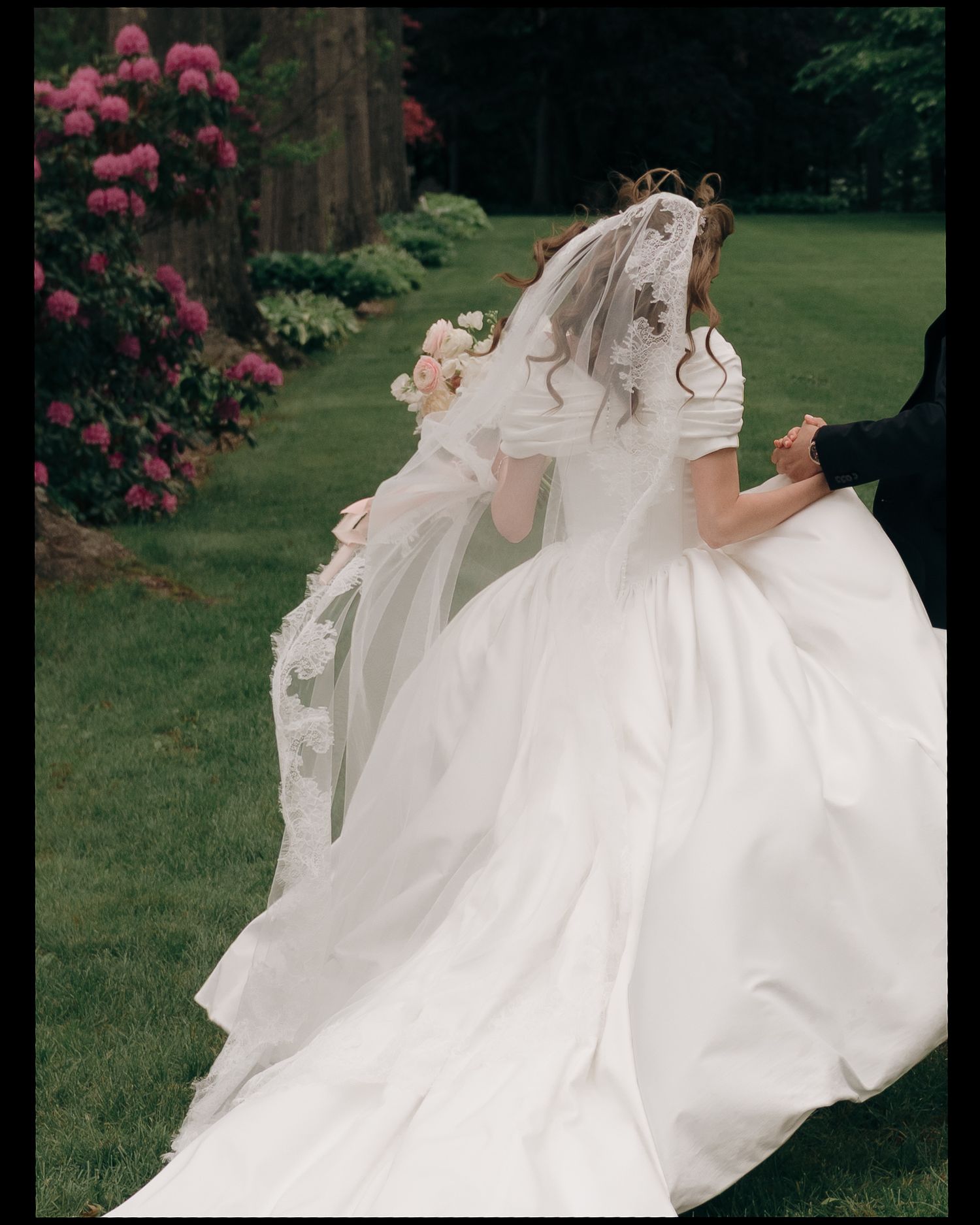 A bride in a flowing white wedding dress twirls outdoors in a garden with blooming pink flowers and green lawn.