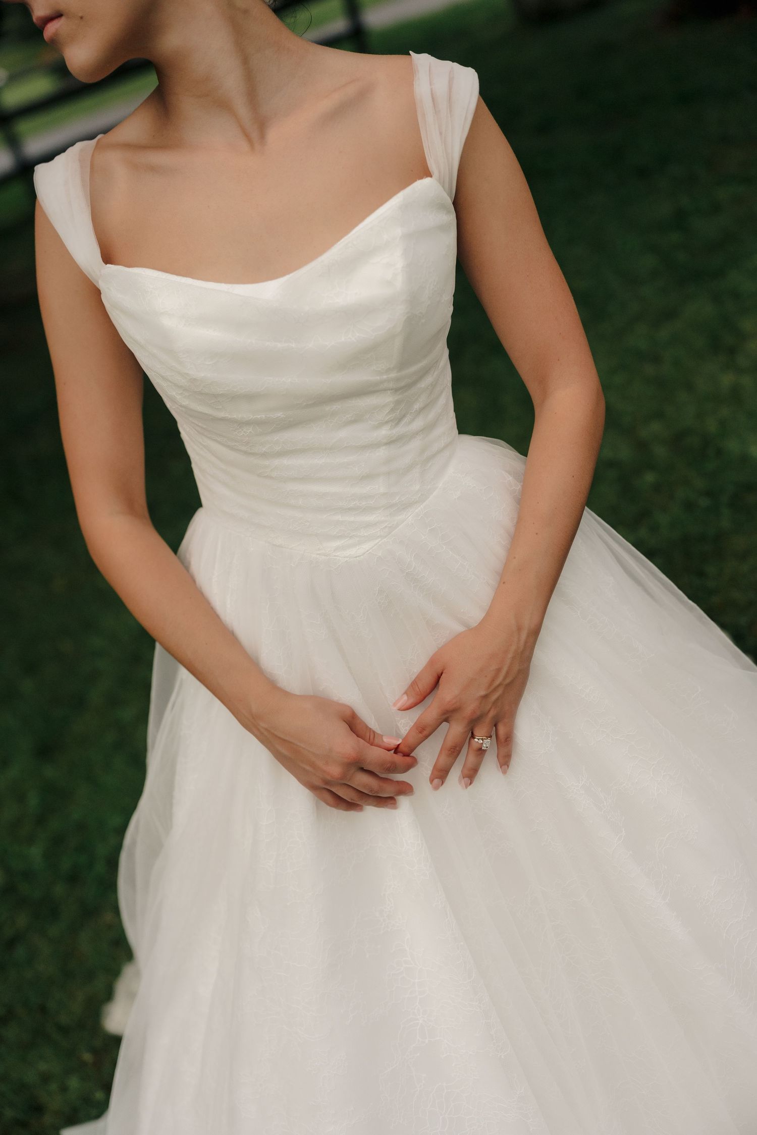 Bride in elegant white tulle ball gown with square neckline and ruched bodice, hands clasped at waist outdoors.