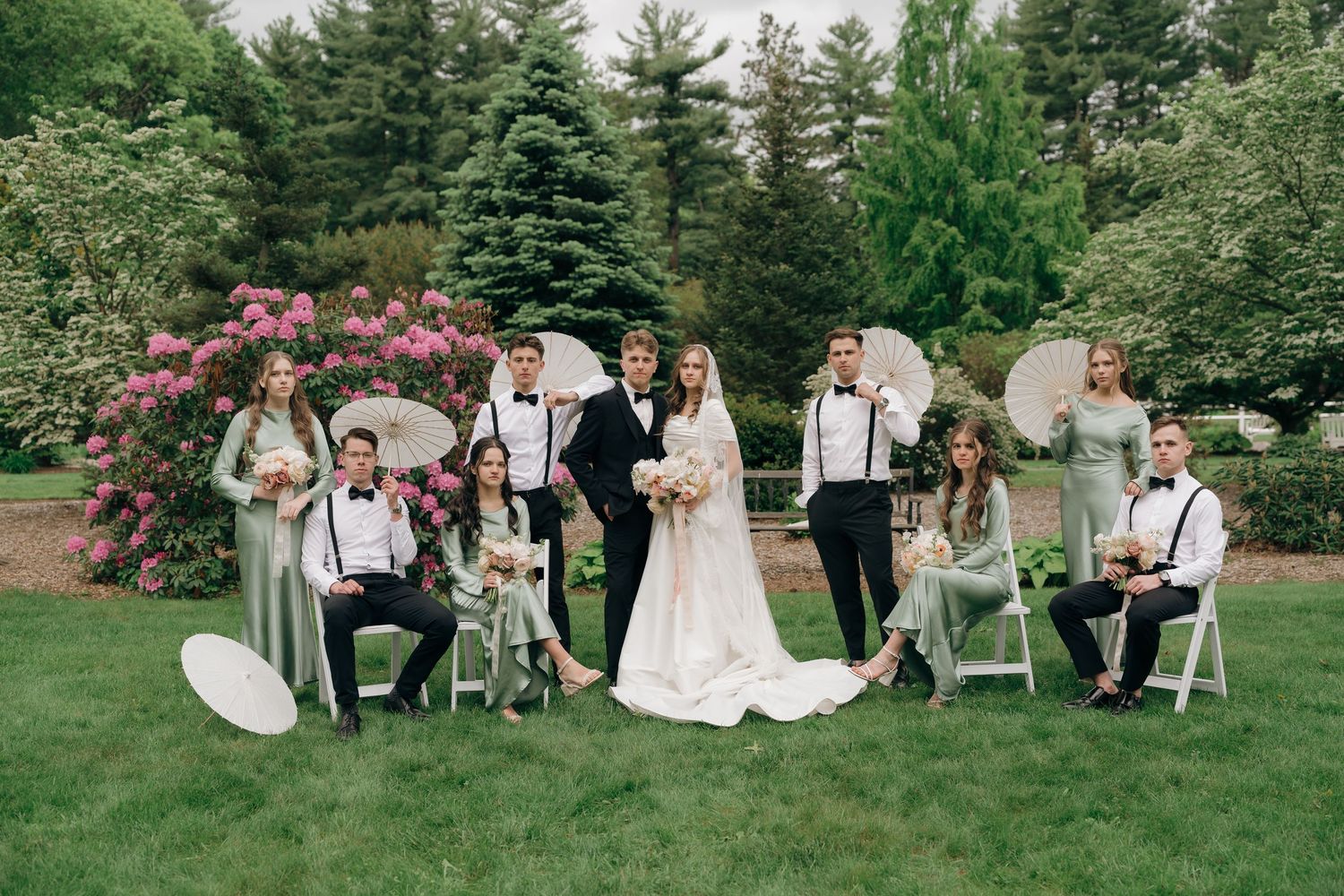 Wedding party relaxes on white chairs arranged outdoors with evergreen trees and pink flowers in the background.