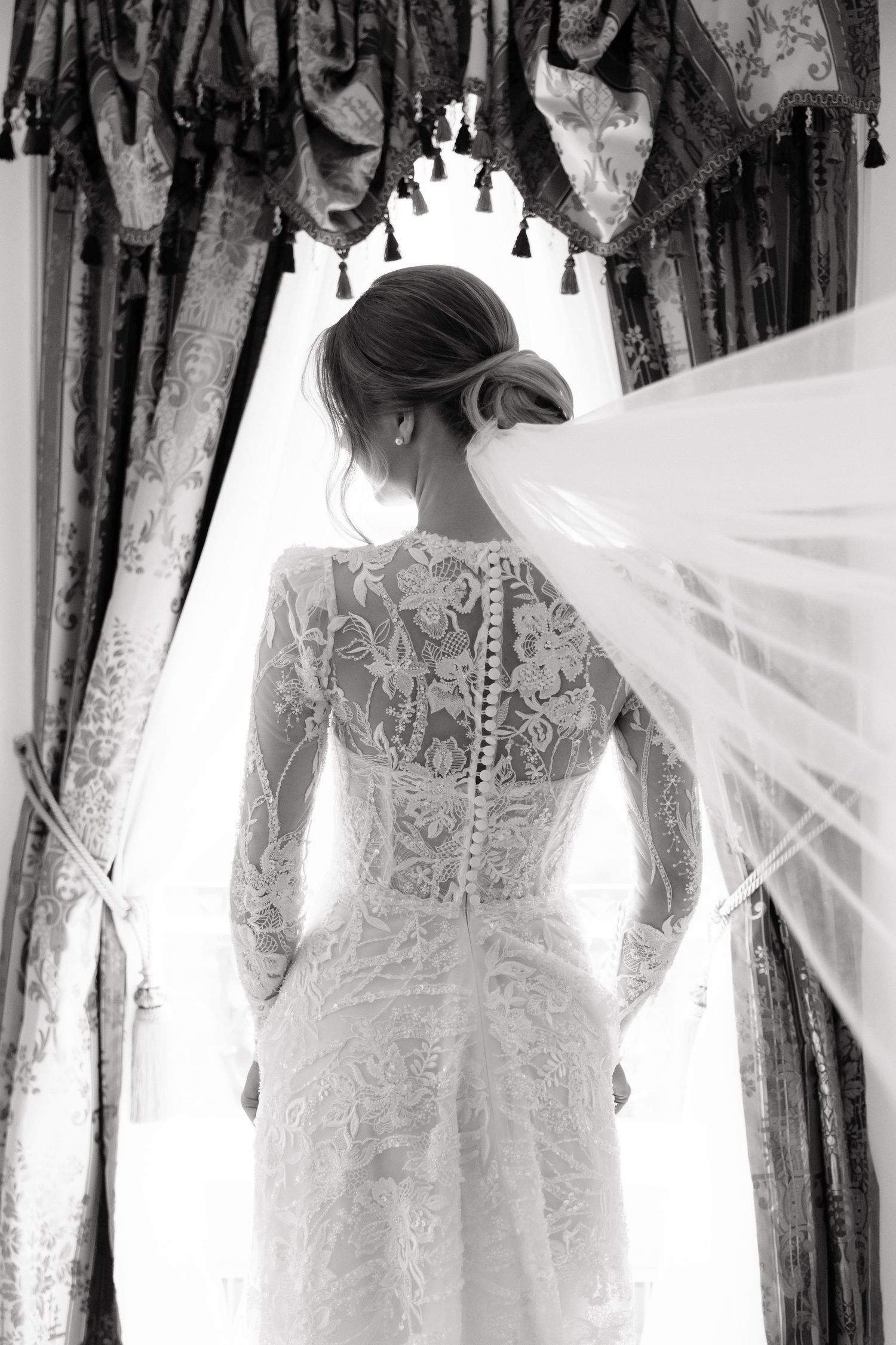 Black and white photo of bride in lace long-sleeve wedding dress with veil flowing, seen from behind at window.
