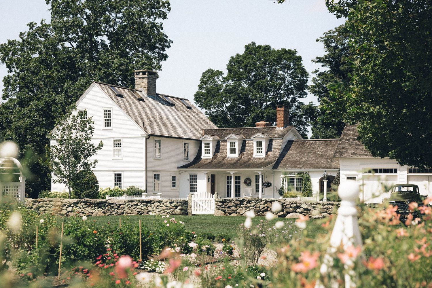 A scenic panoramic view of a white farmhouse with dormers surrounded by lush trees and blooming gardens.
