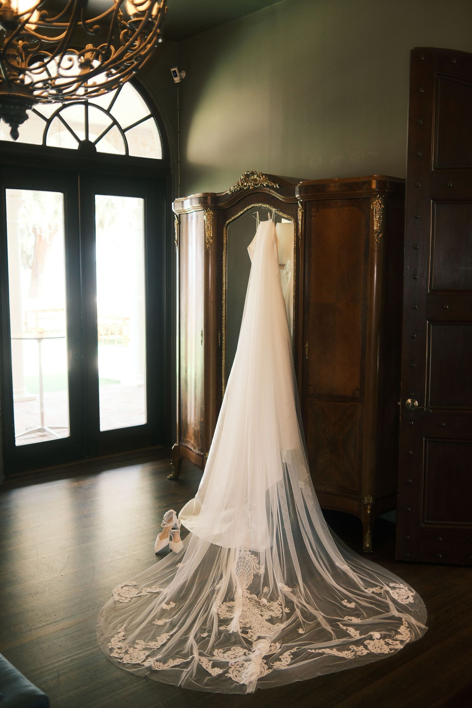 Elegant white wedding dress with long flowing veil displayed on dark hardwood floor with natural lighting.