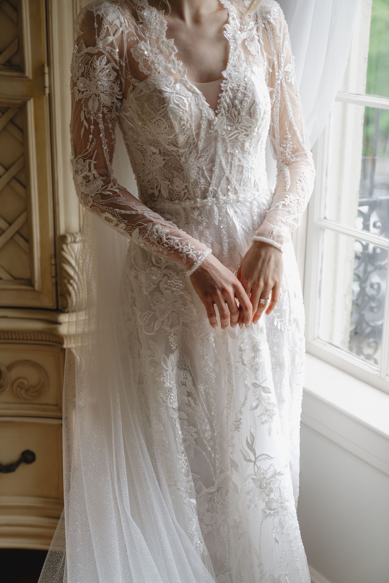 Bride in elegant long-sleeve lace wedding dress standing by window, intricate floral embroidery detail.