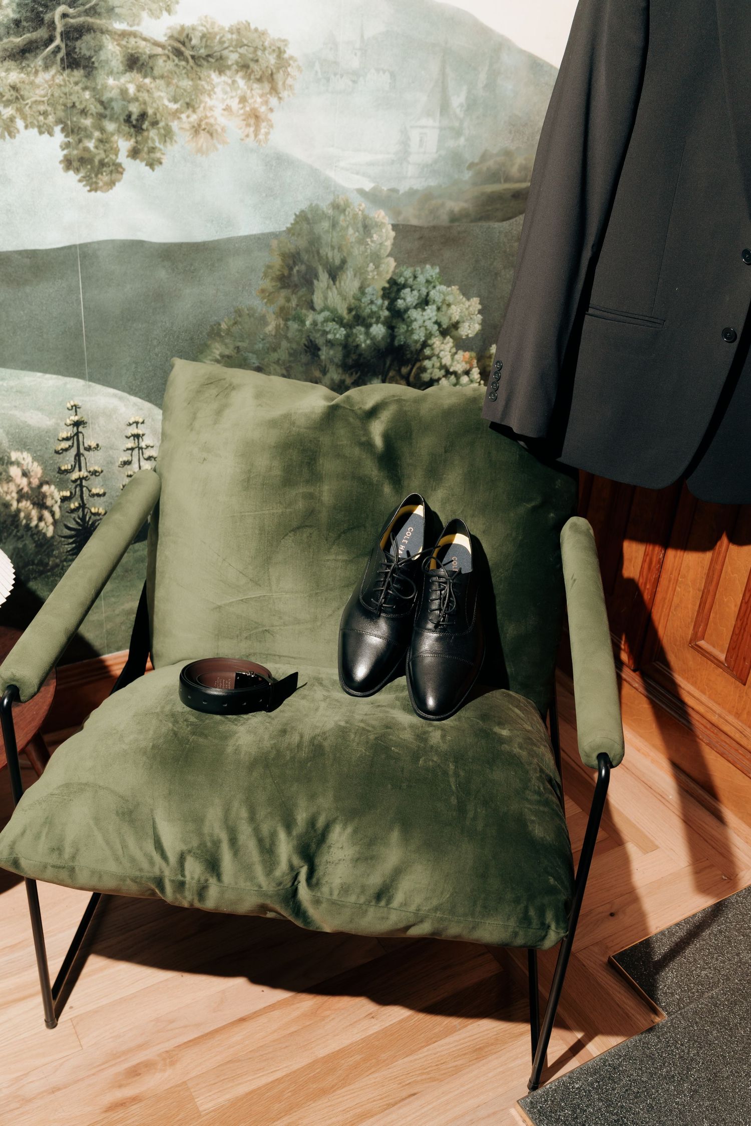 Black dress shoes and belt on green velvet chair with suit jacket hanging nearby, elegant wedding preparation setup.