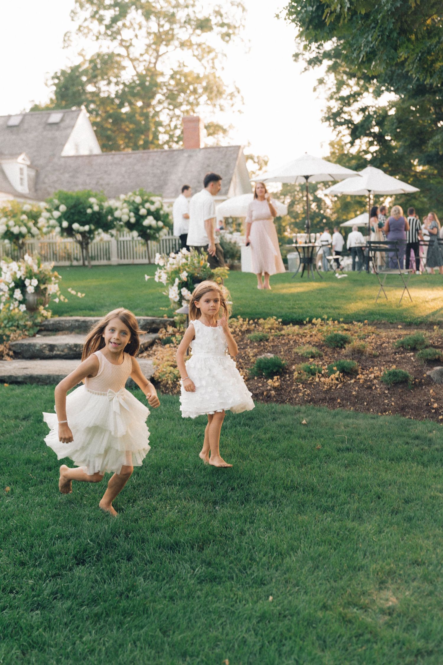Children in white dresses run and play joyfully on a grassy lawn during an outdoor event.