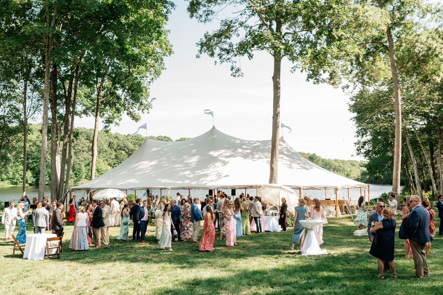 Guests mingle on green lawn beside large white sperry tent at outdoor summer wedding reception surrounded by tall trees.