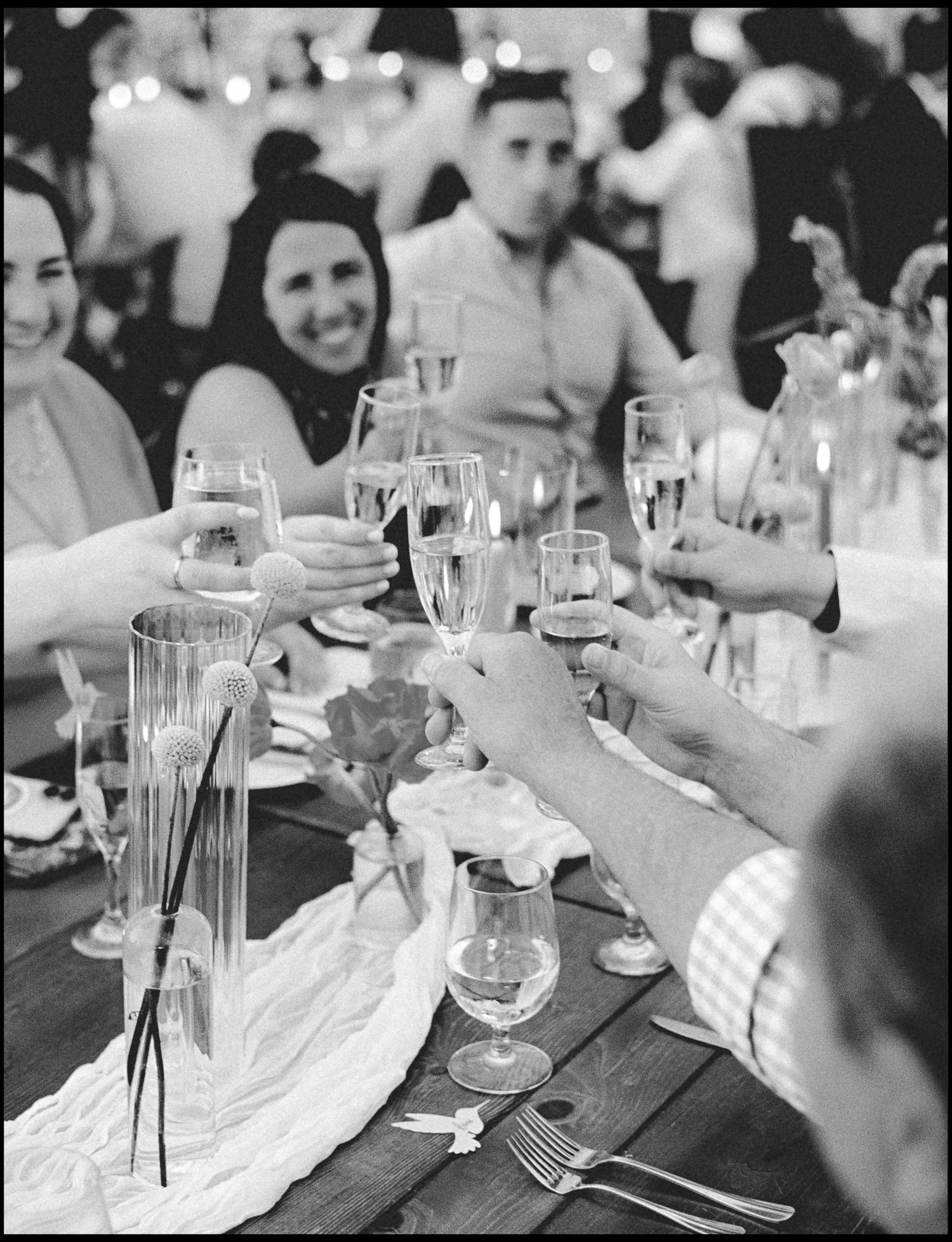 Black and white photo of wedding guests clinking champagne glasses in a toast at a decorated reception table.
