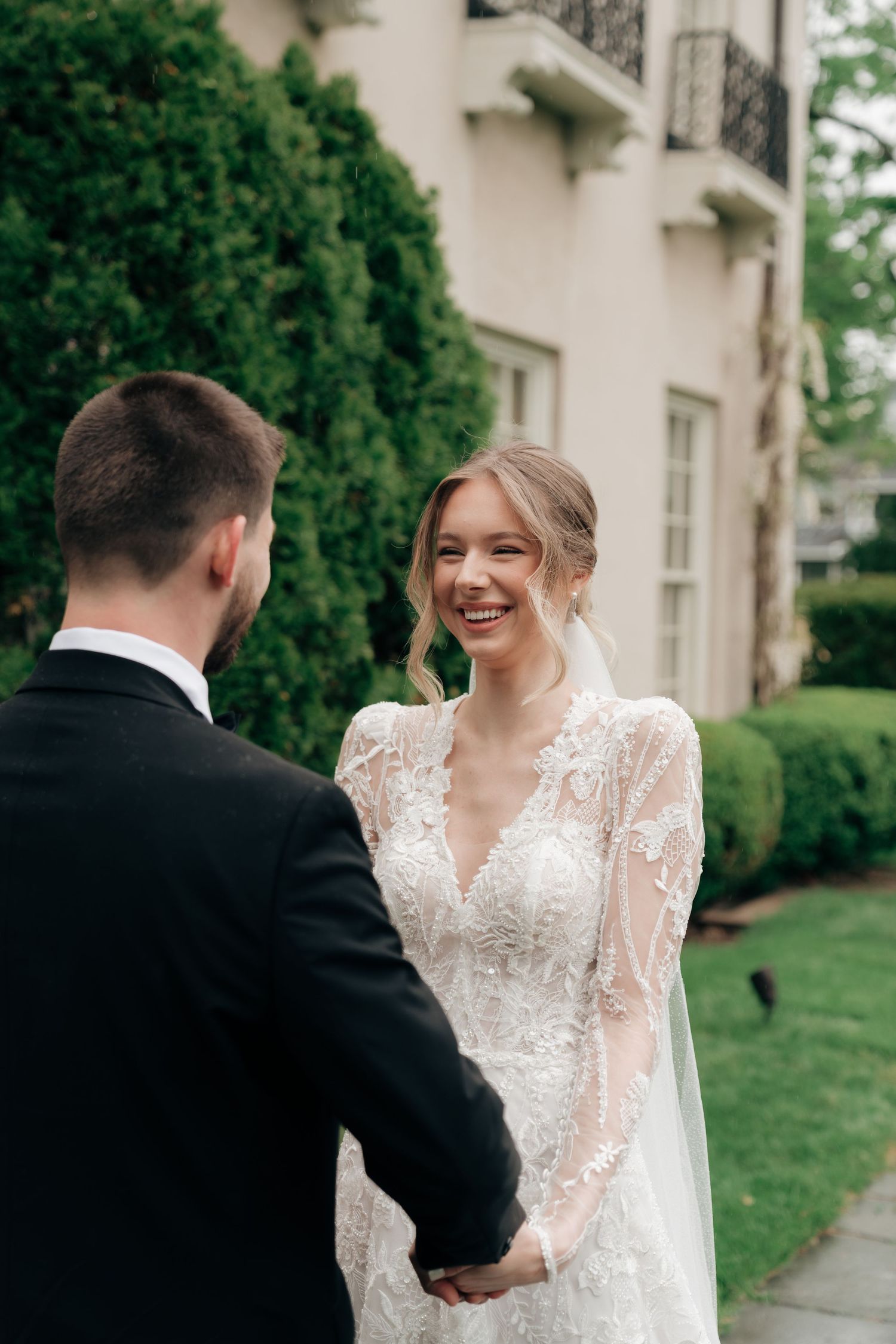 A smiling bride in a lace wedding gown holds hands with her groom in a black suit outdoors near a elegant building.