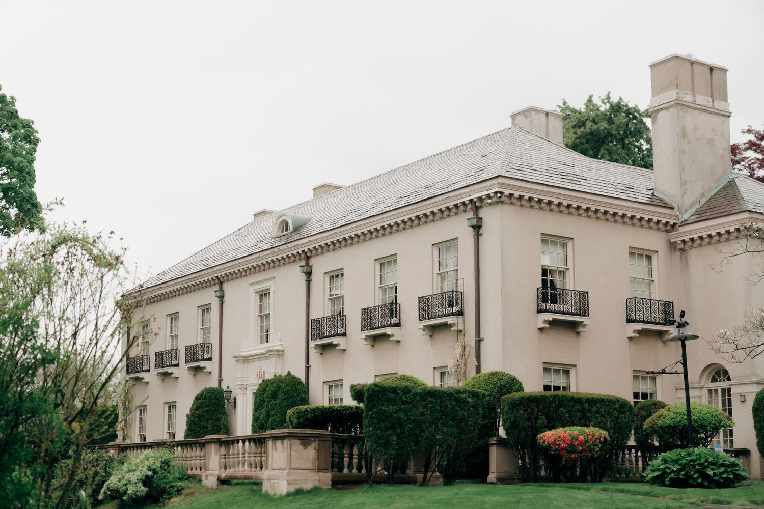 Elegant neoclassical mansion with balconies and manicured hedges surrounded by green trees.