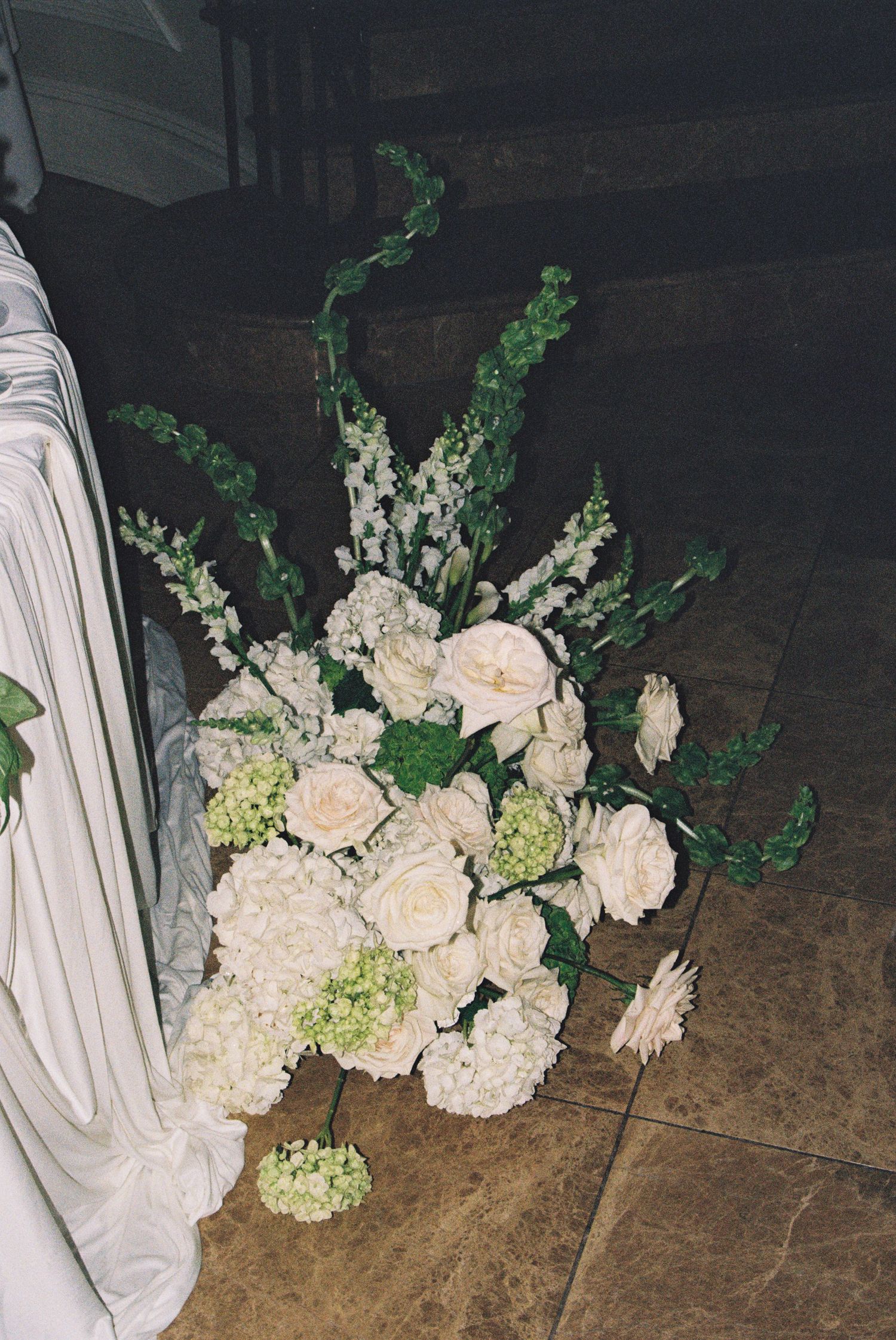 Elegant white floral arrangement with roses and hydrangeas on a tiled floor beside draped fabric at a wedding ceremony.