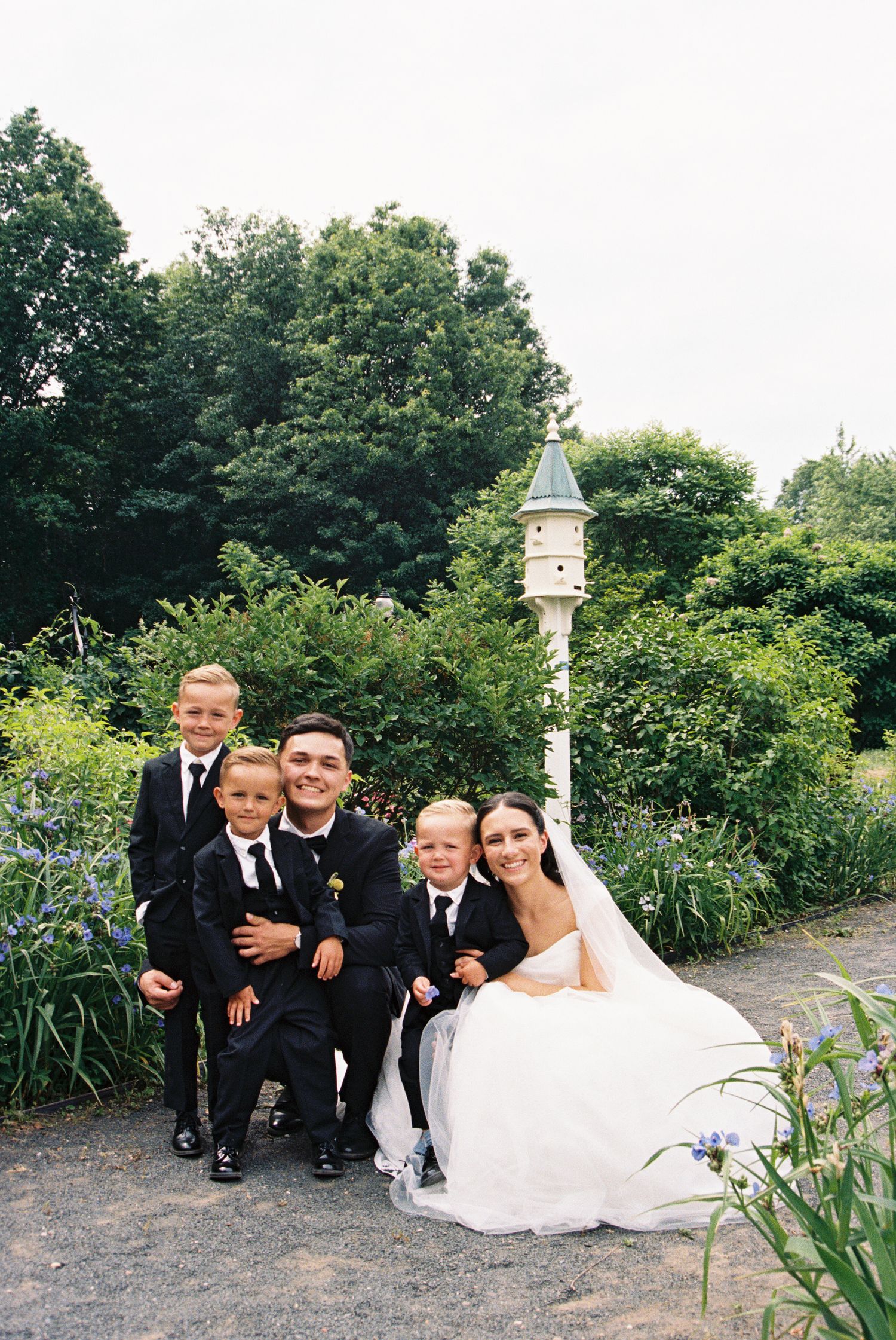 Bride and groom pose with three boys in black suits in a lush garden setting with a white birdhouse.