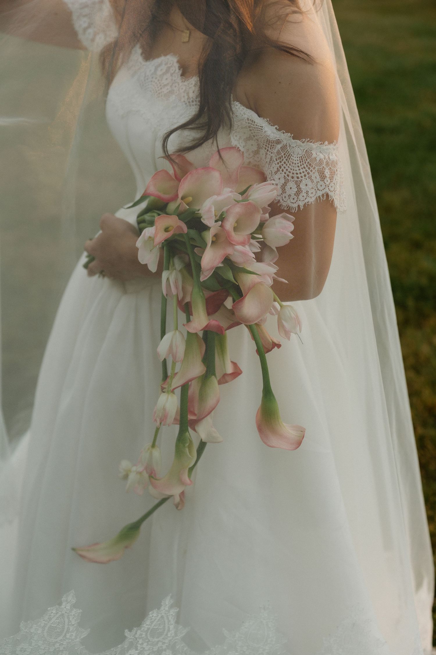 Bride in white off-shoulder lace gown holding cascading pink calla lily and tulip bouquet outdoors.