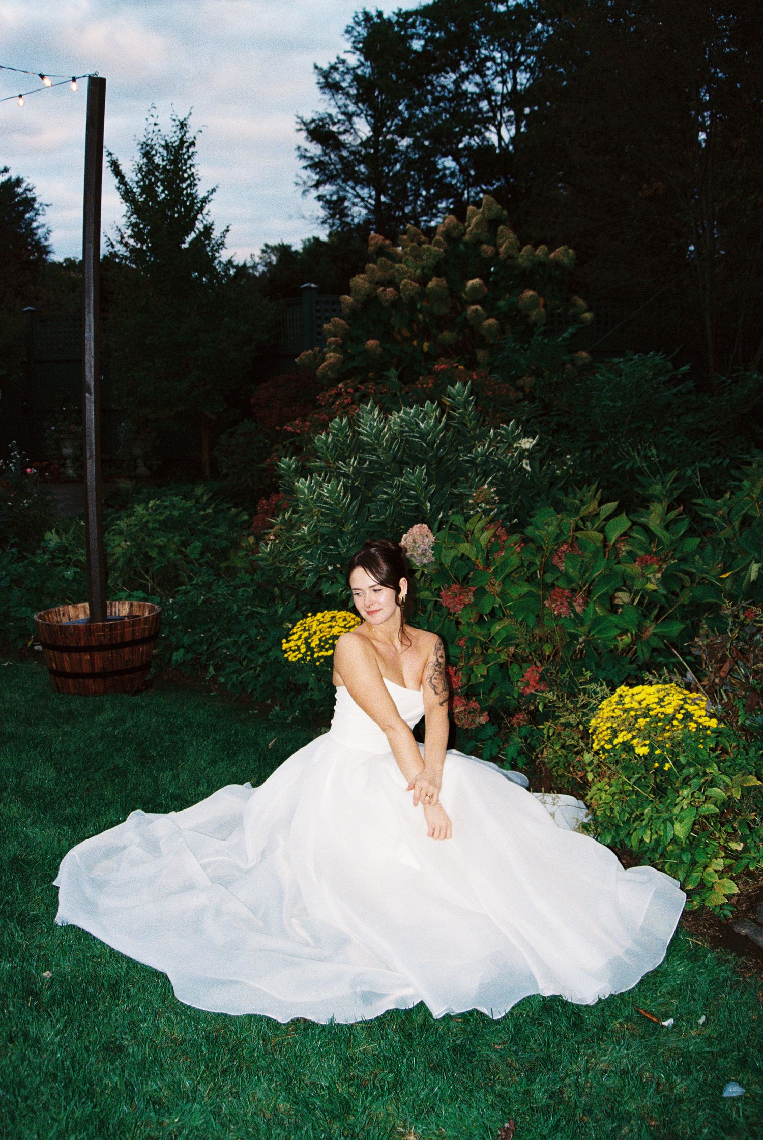 Bride in flowing white gown poses gracefully on garden lawn with yellow flowers.