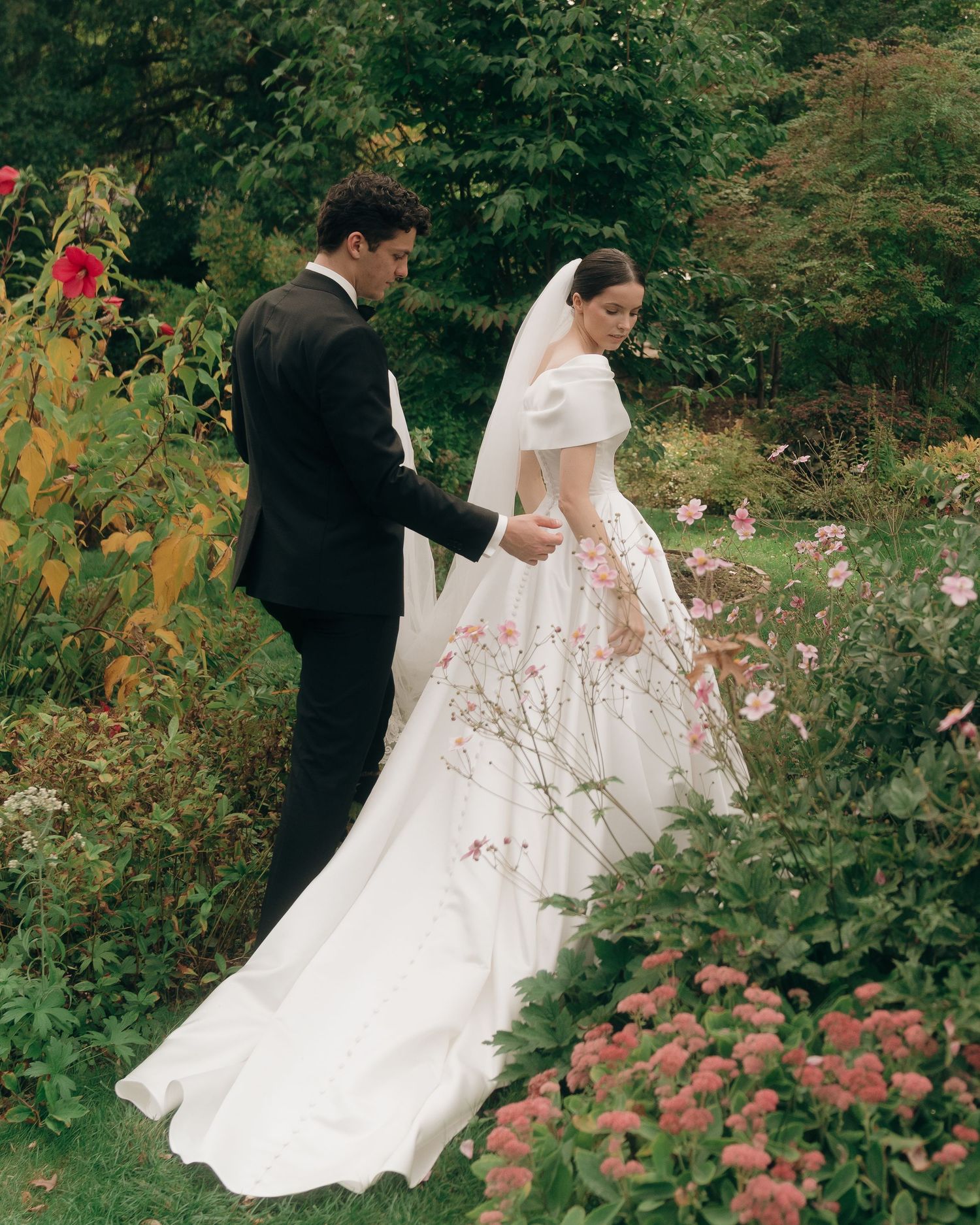 A couple in wedding attire strolls through a lush garden filled with blooming pink roses and greenery.