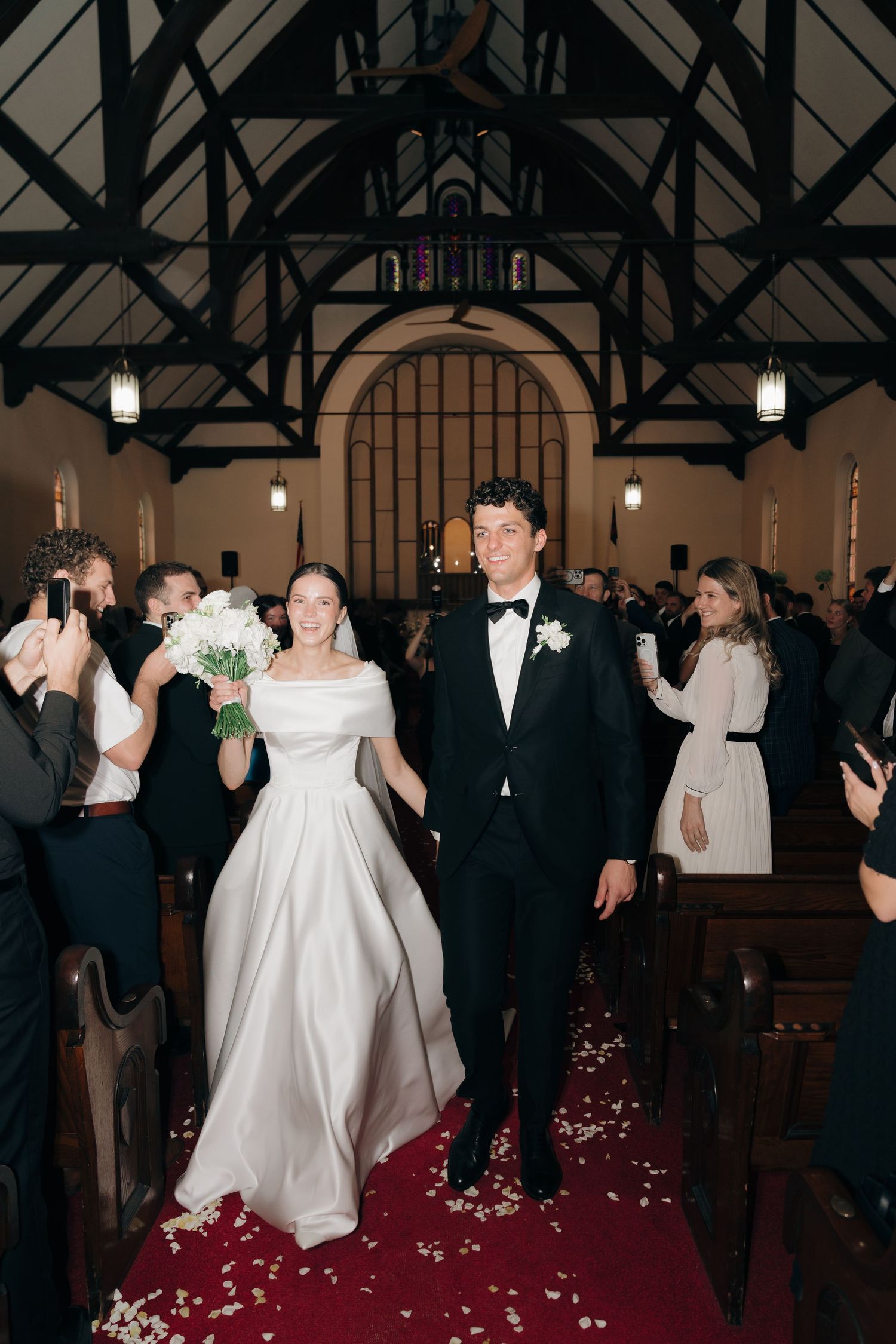 Wedding party walks down red-carpeted aisle in church with rose petals.