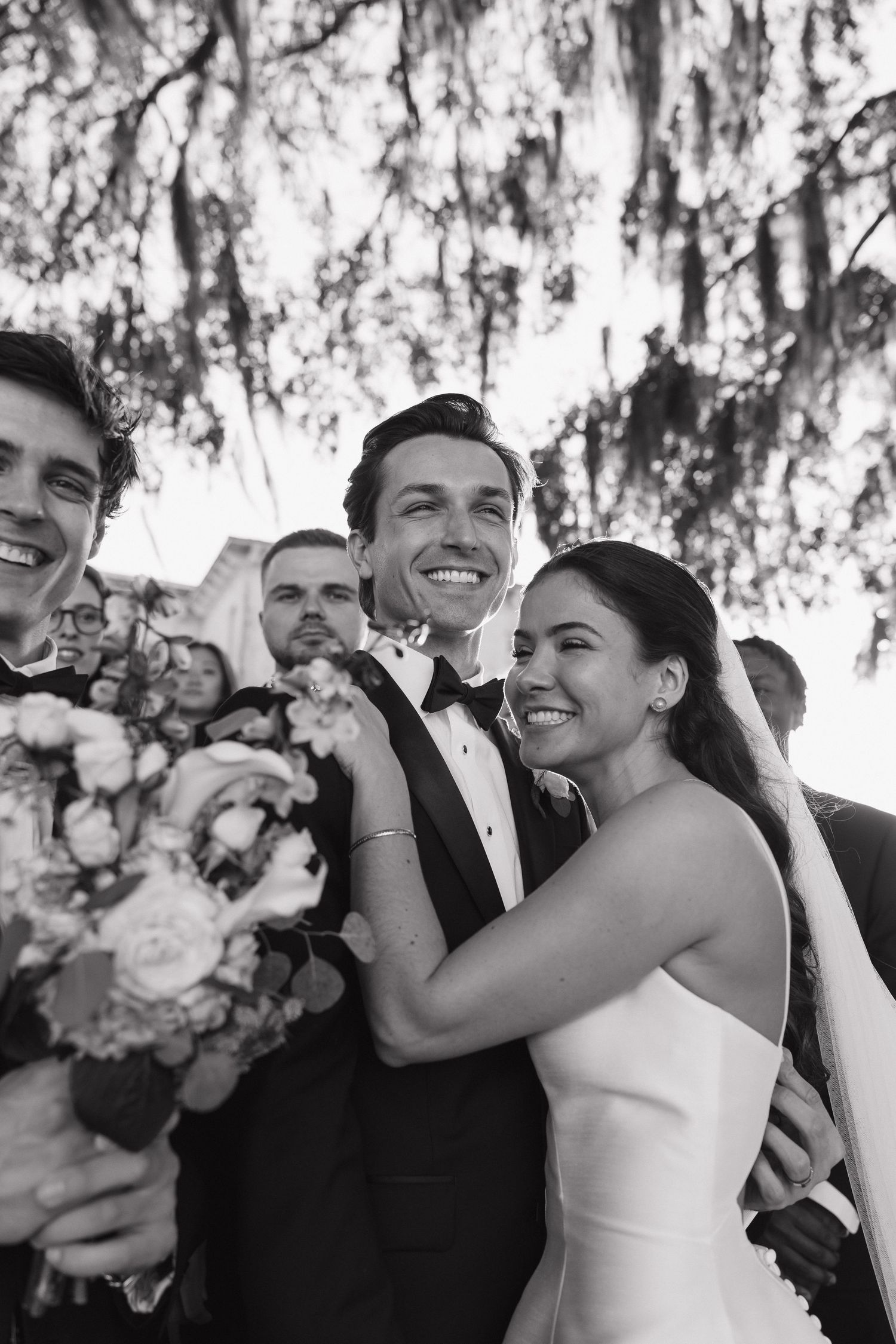 Wedding party members laugh joyfully together outdoors under trees with white floral bouquets.