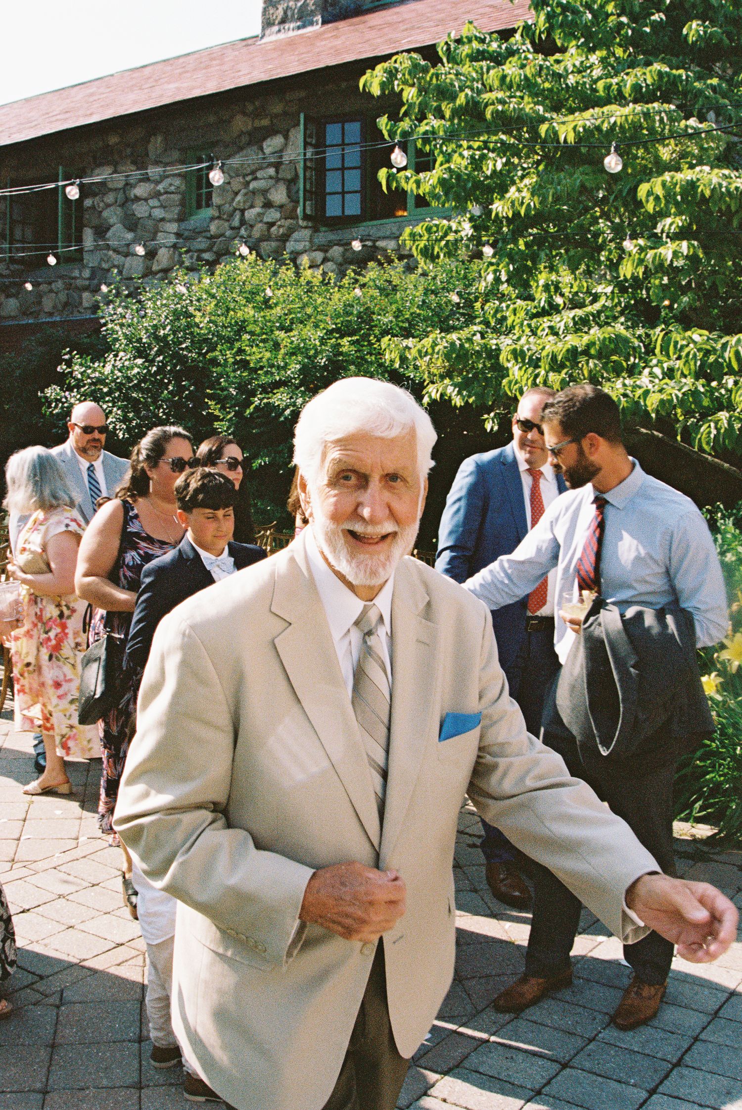 Elderly man in beige suit with blue pocket square smiles at outdoor wedding reception near stone building.