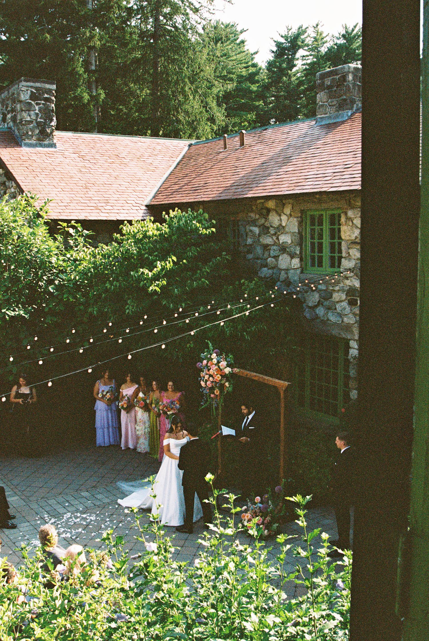 Outdoor wedding ceremony in a stone courtyard with string lights, floral arch, bride and groom, and bridesmaids in pastel gowns.
