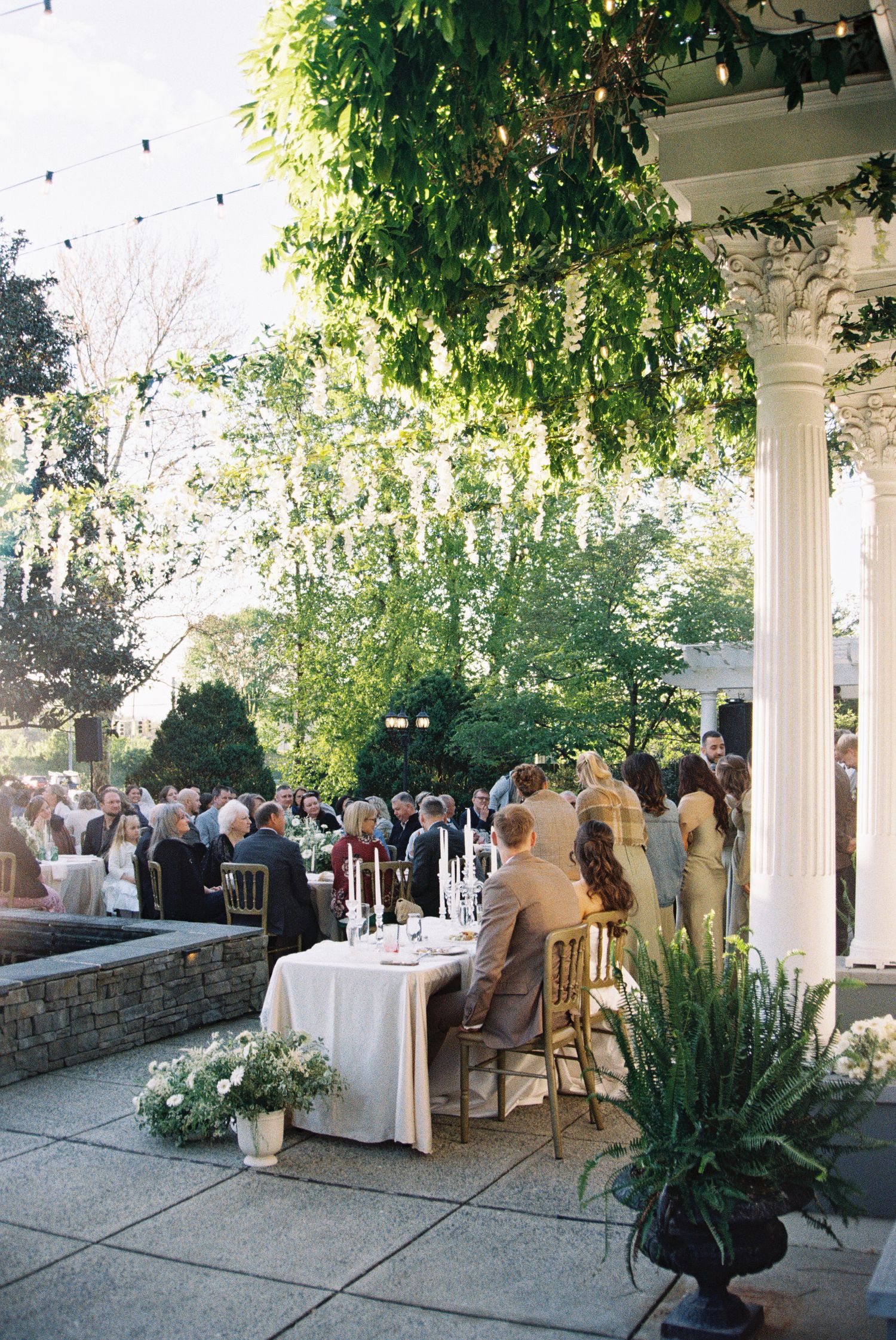 Elegant outdoor wedding reception under lush greenery with string lights, white columns, and guests seated at candlelit tables.