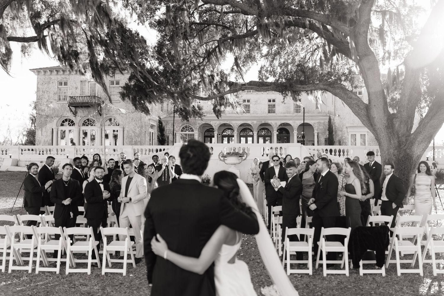 A black and white panoramic collage of an outdoor wedding ceremony with guests seated under large trees.