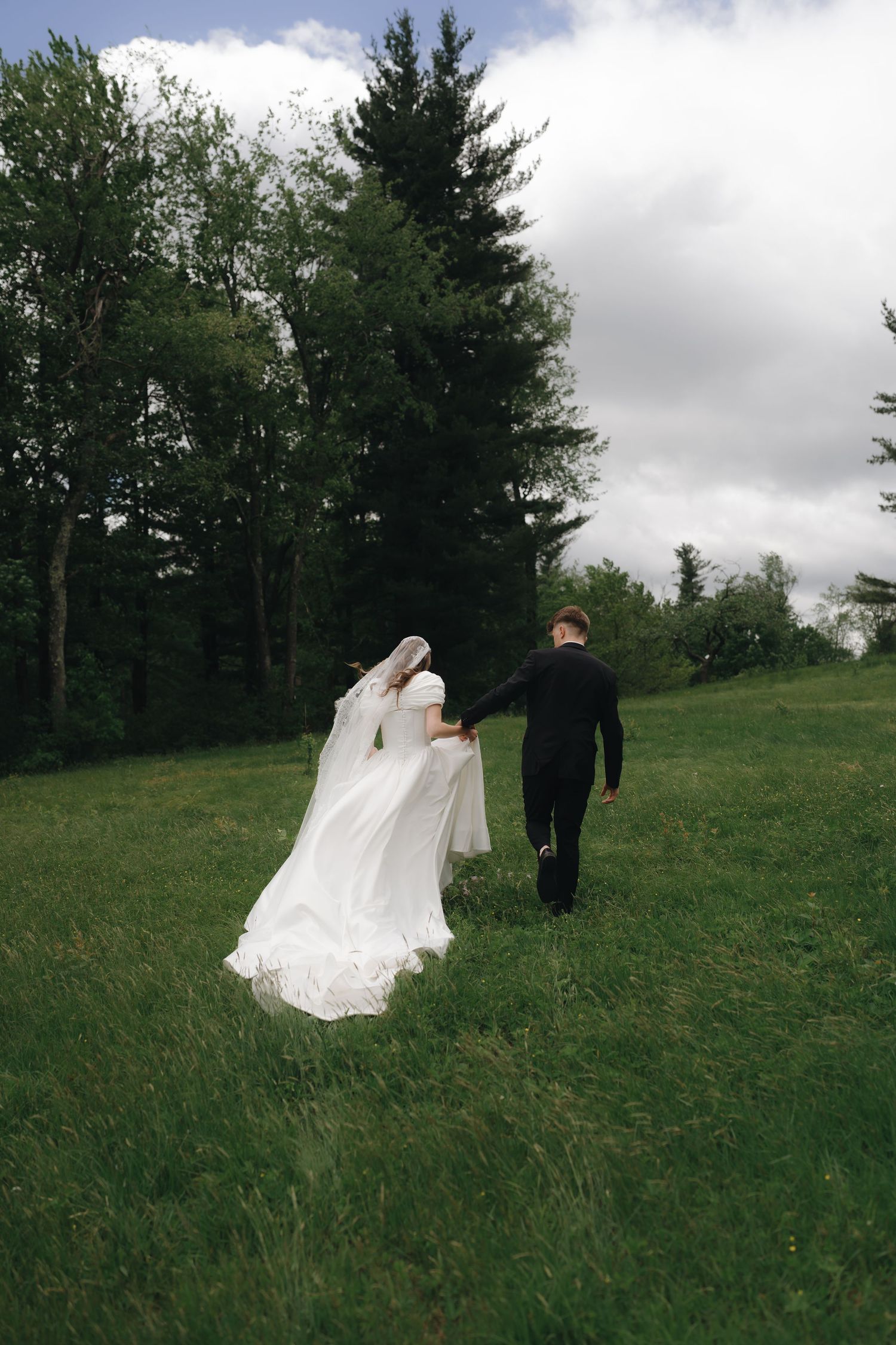 A wedding couple walks hand in hand up a grassy hill against a cloudy sky and pine tree backdrop.