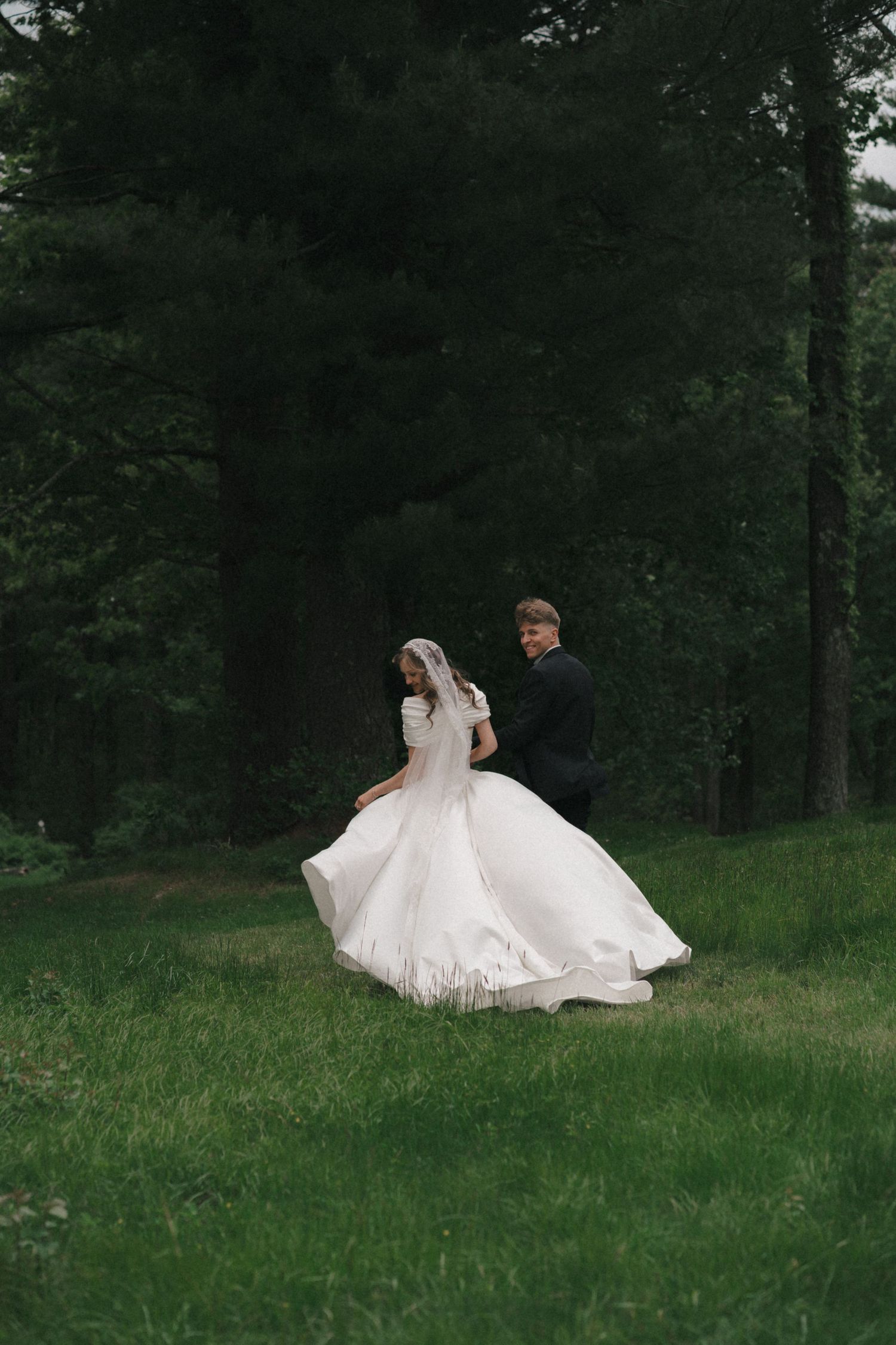 A couple in wedding attire strolls across a green hillside surrounded by evergreen trees.