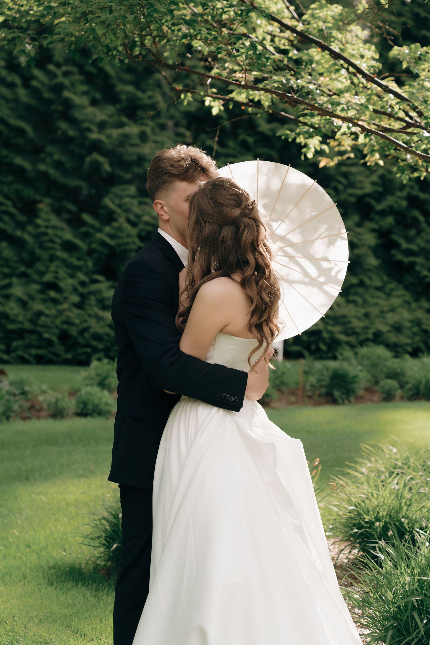 A couple in wedding attire poses in a garden setting with a white parasol creating a romantic outdoor scene.