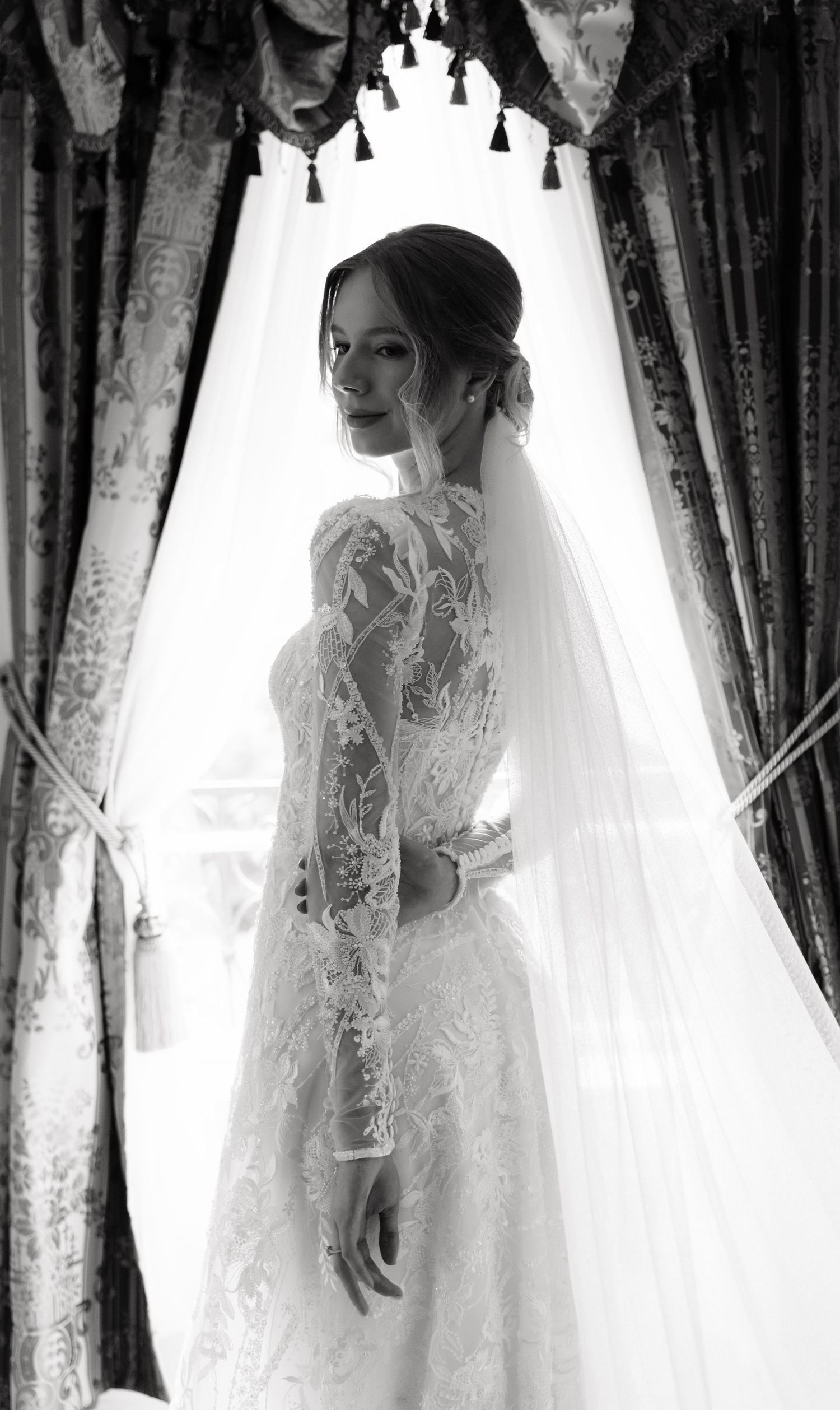 Elegant bride in lace long-sleeve wedding gown and veil stands by draped curtains in black and white portrait.