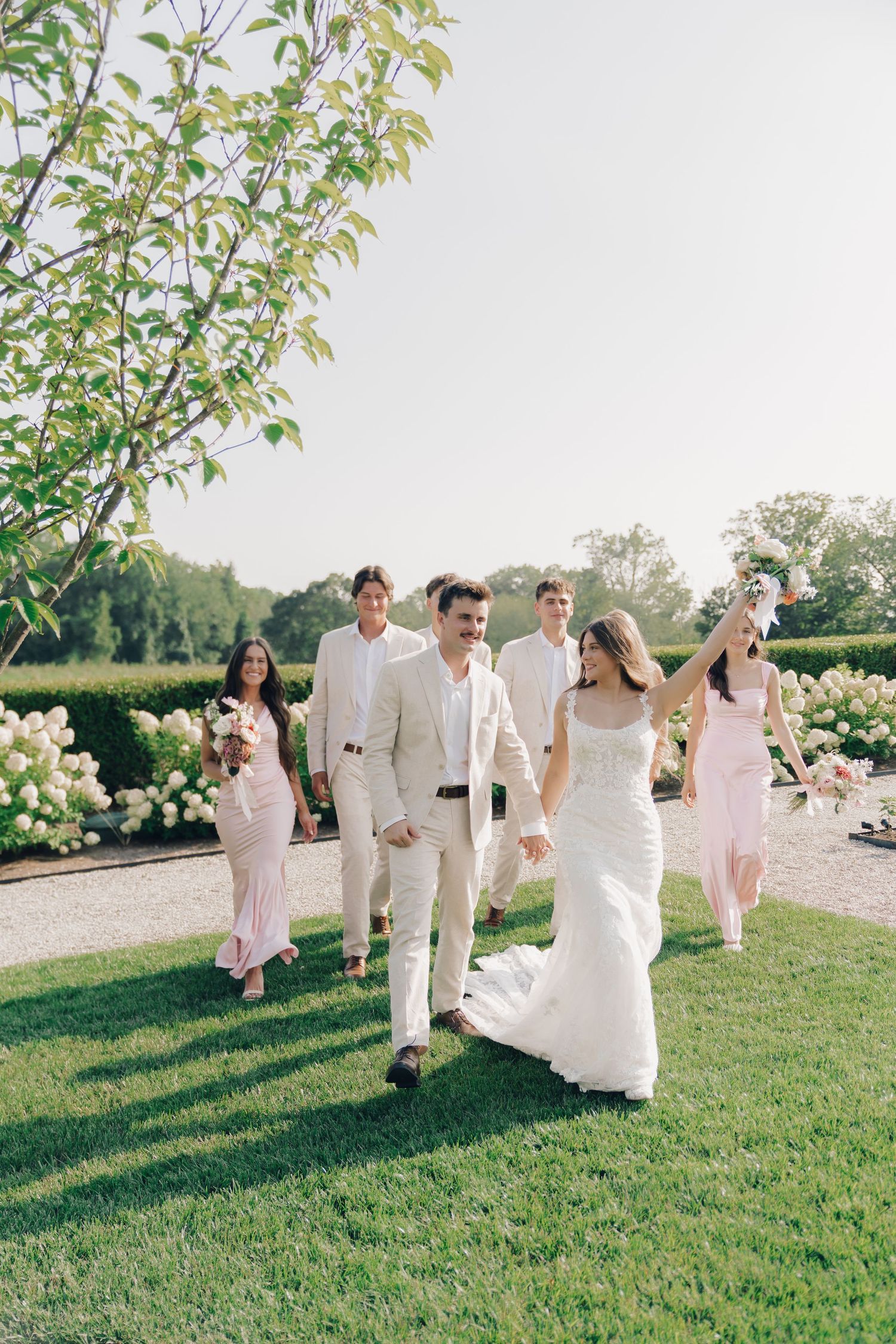 Wedding party in pink and white attire dancing and celebrating on a lush green lawn at an outdoor venue.