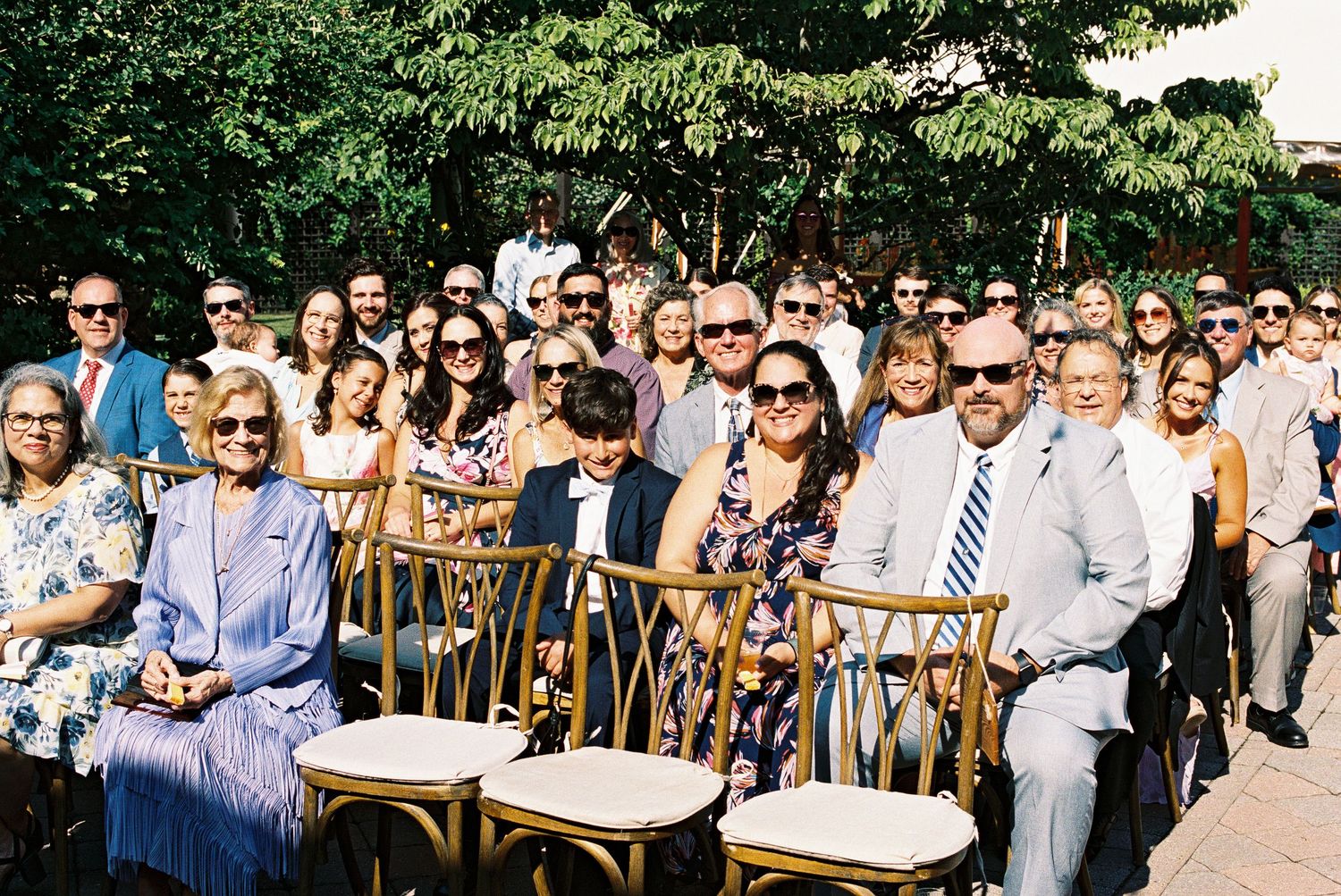 Smiling wedding guests seated outdoors in gold chairs at a sunny ceremony, dressed in elegant summer attire.