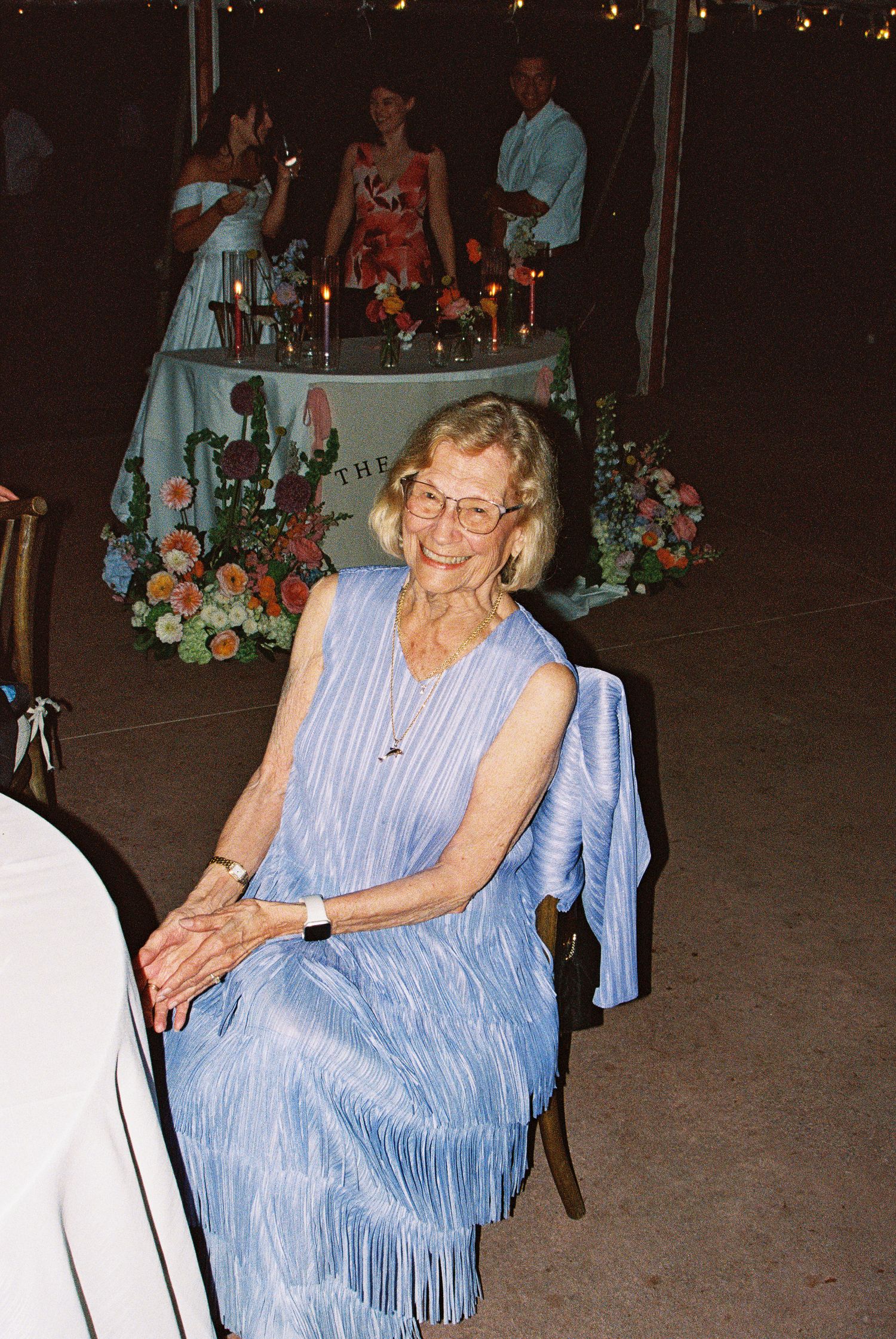 Elderly woman in blue fringed dress smiling at outdoor party with floral decorations and candles in background.