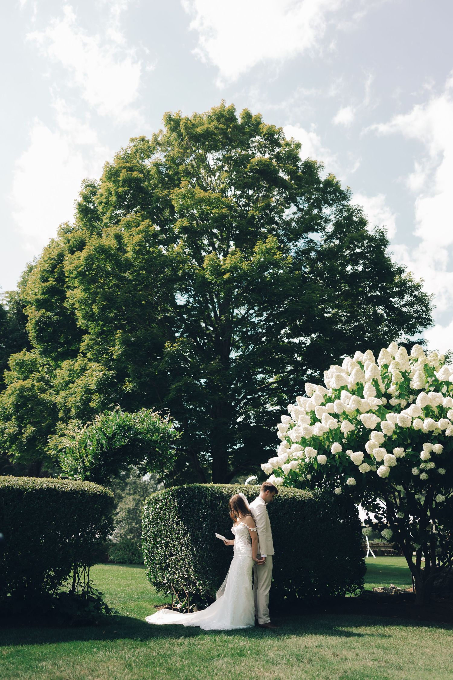 A couple shares a romantic moment in a garden with lush trees and blooming white hydrangeas on their wedding day.